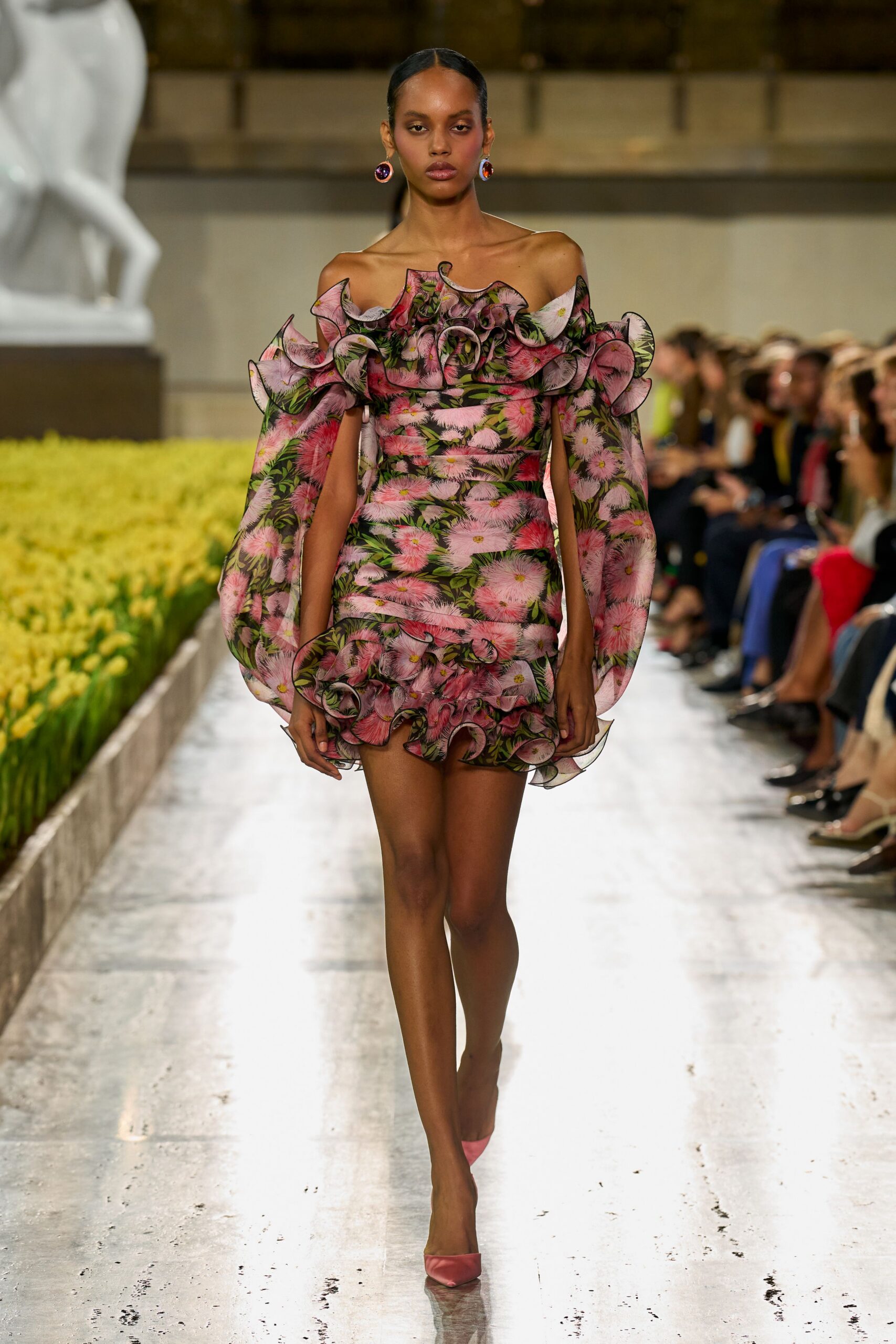 Off-shoulder floral mini in sheer organza, wired ruffles curling at neck and hem. Detached balloon sleeves in the same pink blossom print. Round cabochon drop earrings, plum and amber glass. Petal-pink satin pumps with a sharp vamp. Walking beside formal flowerbeds just after the sprinklers clicked off.