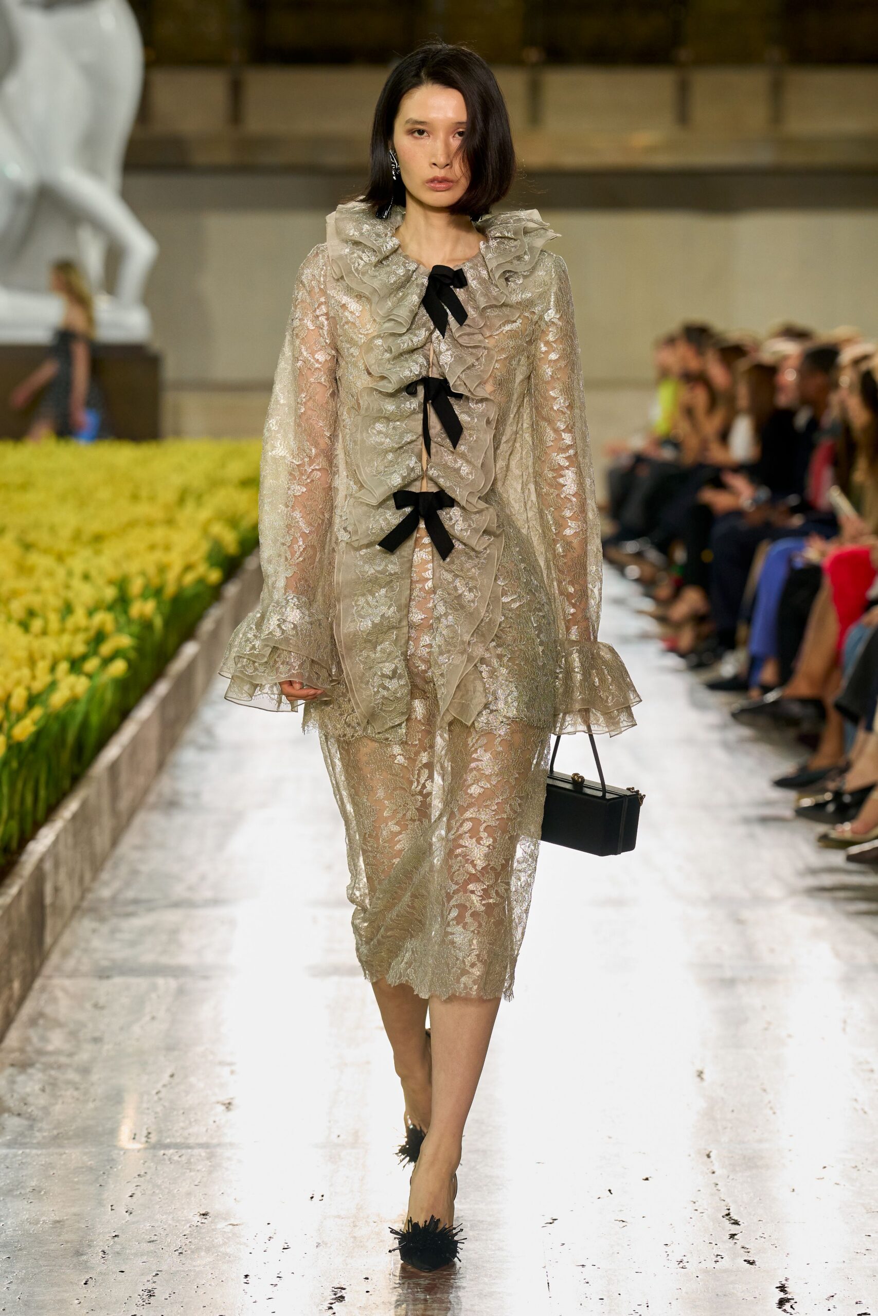 Sheer champagne lace dress with a deep ruffled placket tied in black bows. Boxy black top-handle bag with tiny gold ball hardware. Black pointed pumps tipped with spiky feather tufts. Crossing a sculpture court after the wedding, shoes catching on fallen petals.