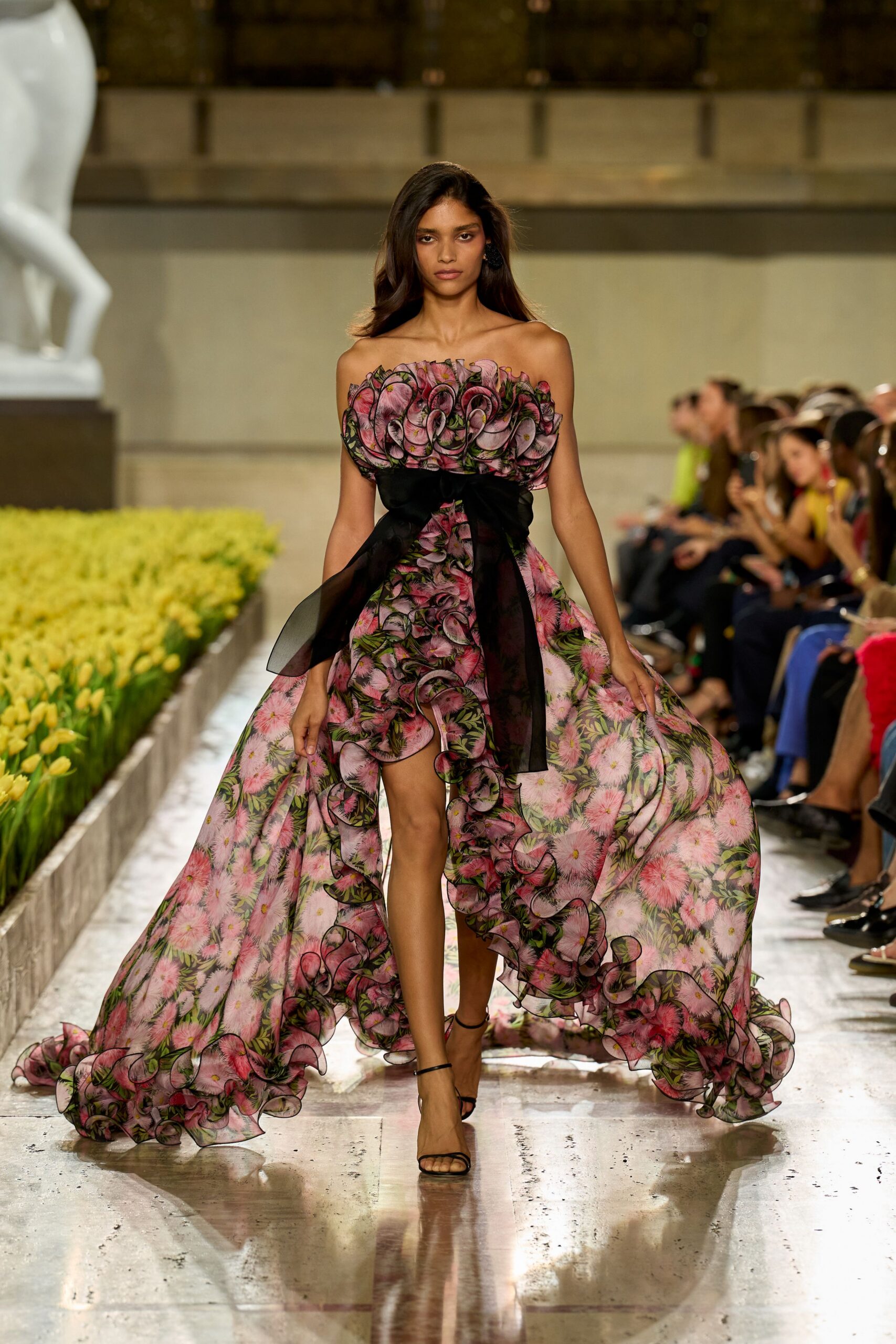 Strapless floral organza gown with looped black-edged ruffles and a high-low train. Wide black sheer sash knotted hard at the waist. Barely there black ankle-strap sandals. Crossing a conservatory aisle while the flower delivery is still being unpacked.