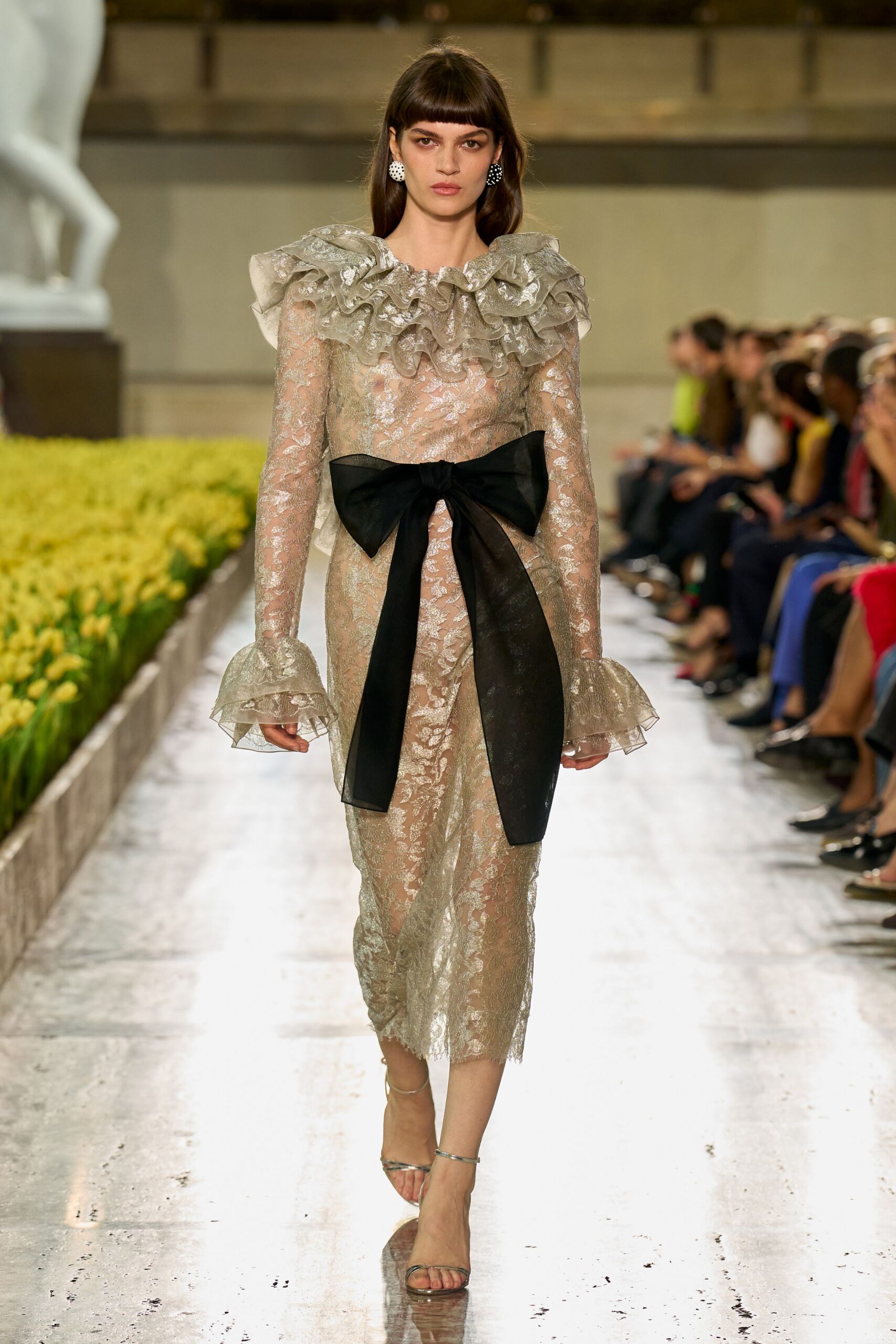 Round black-and-white dot earrings. Sheer silver lace column dress with stacked organza ruffles at neck and cuffs. Oversized black bow tied flat across the waist, tails falling past the knee. Barely there metallic ankle-strap sandals. Crossing a museum atrium after the wedding crowd has gone upstairs.