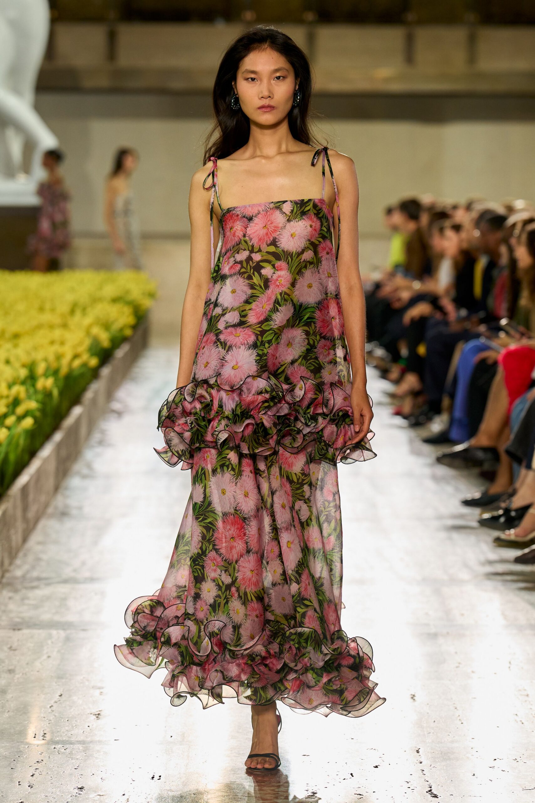 Sheer black floral chiffon slipdress, tied shoulder straps and horsehair ruffles at hip and hem. Blue clustered drop earrings. Black barely-there strap sandals. Crossing a conservatory aisle while the flowers are being watered.