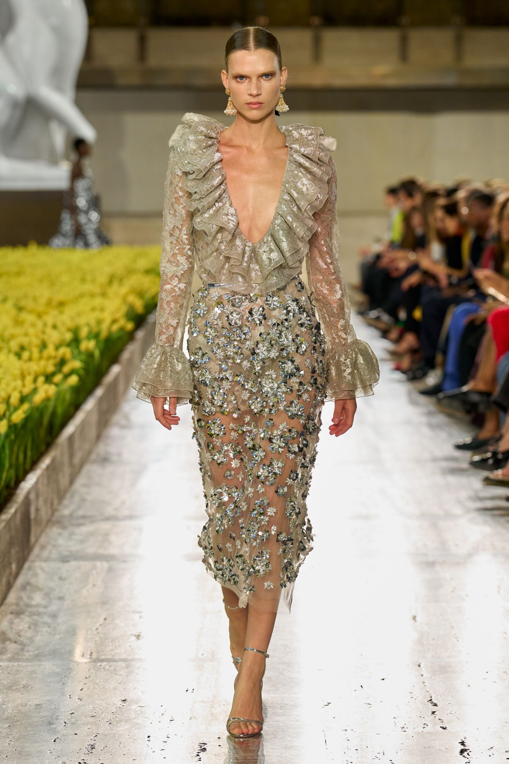 Silver-beige lace gown with a plunging ruffled neckline and flounced cuffs. Sheer column skirt densely appliqued with mirrored flowers over nude lining. Cluster drop earrings. Bare silver ankle-strap sandals. Crossing a conservatory aisle while the last light catches every petal.