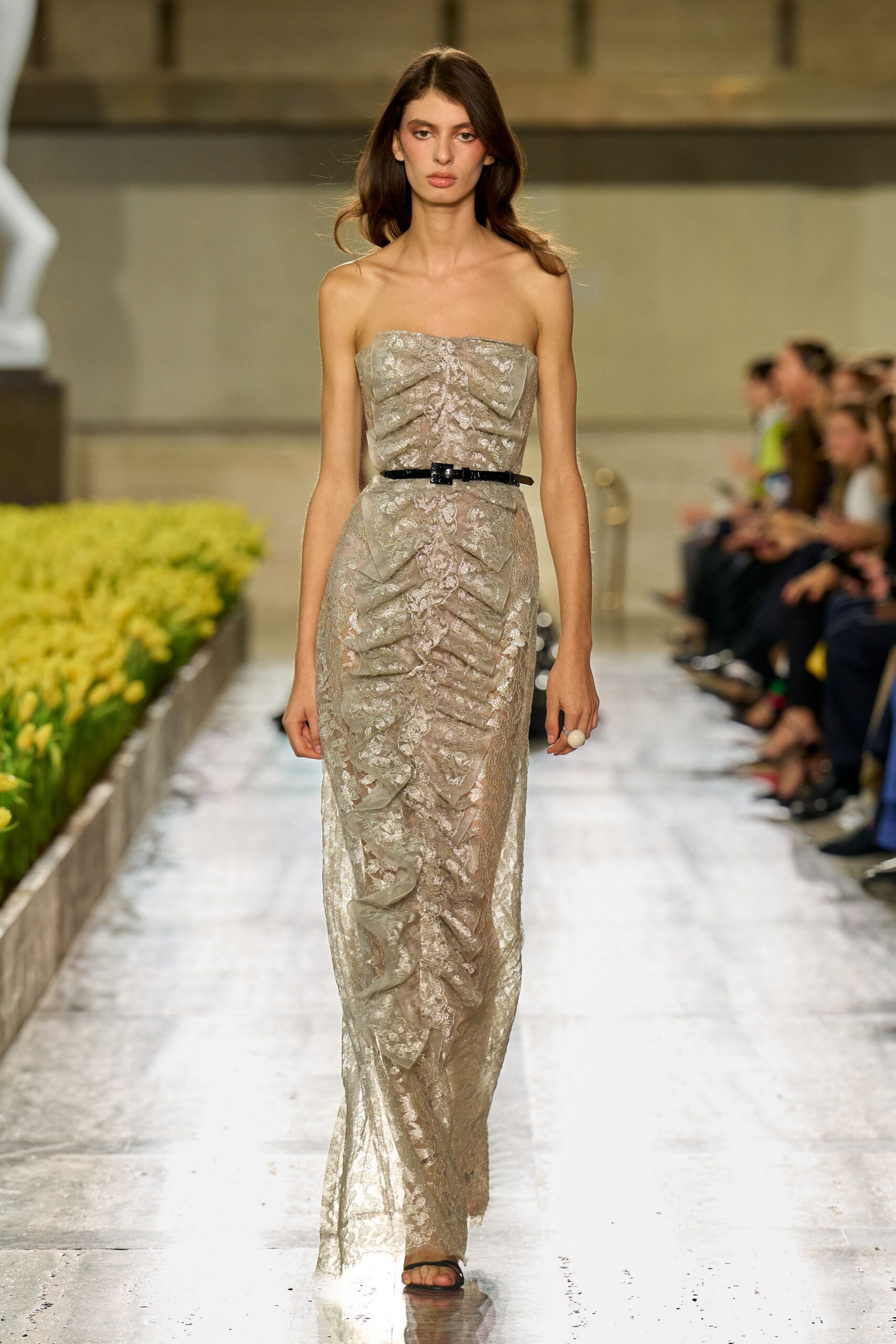 Strapless silver lace column gown, ruched with cascading center ruffles. Thin black patent belt, tiny buckle catching the waist. Large milky cabochon ring. Black barely-there thong sandals. Crossing a glassy conservatory aisle while the flowers are still being watered.