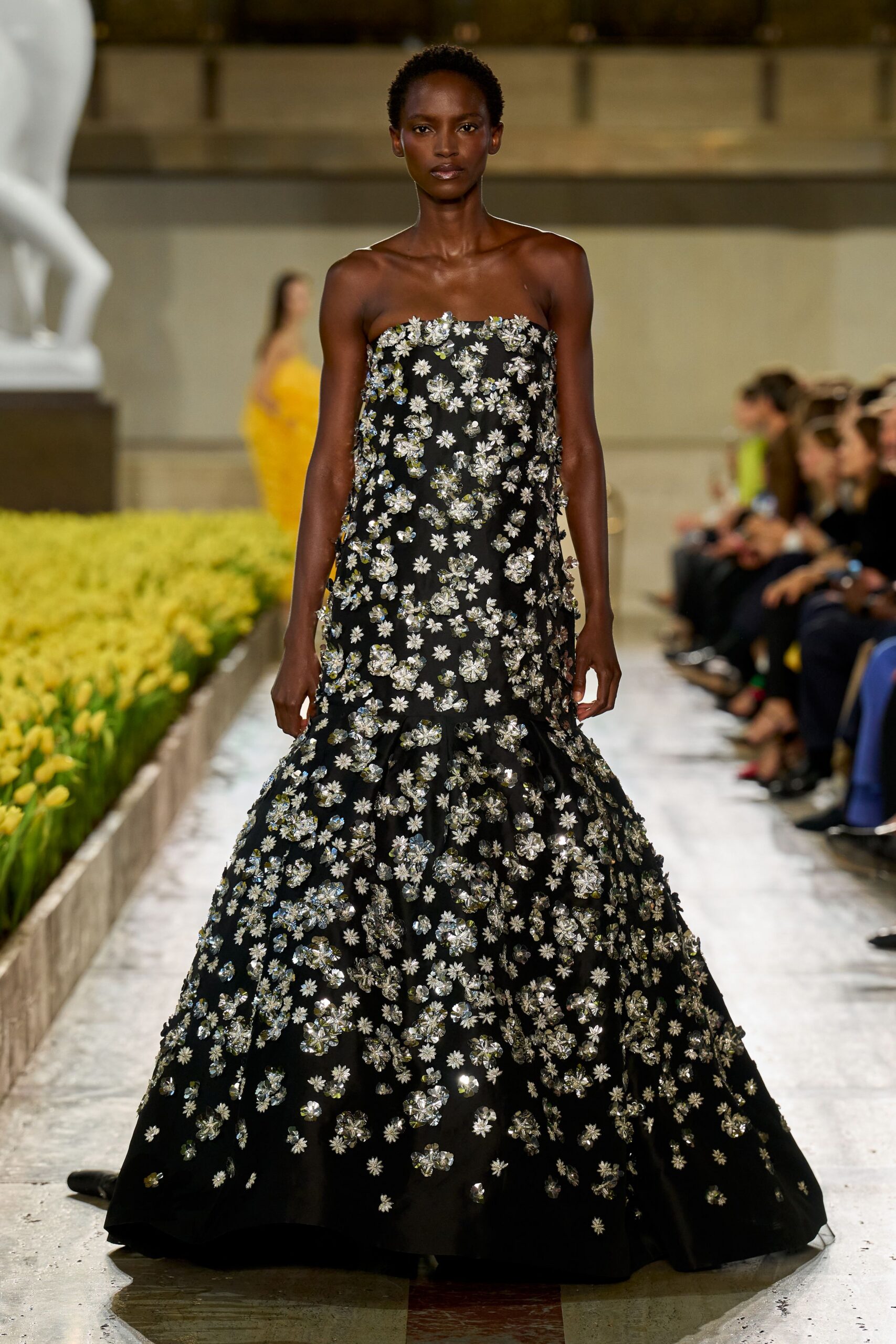 Strapless black ball gown scattered with silver floral appliques and mirrored petals. Crossing a museum atrium while the flower display is being watered.