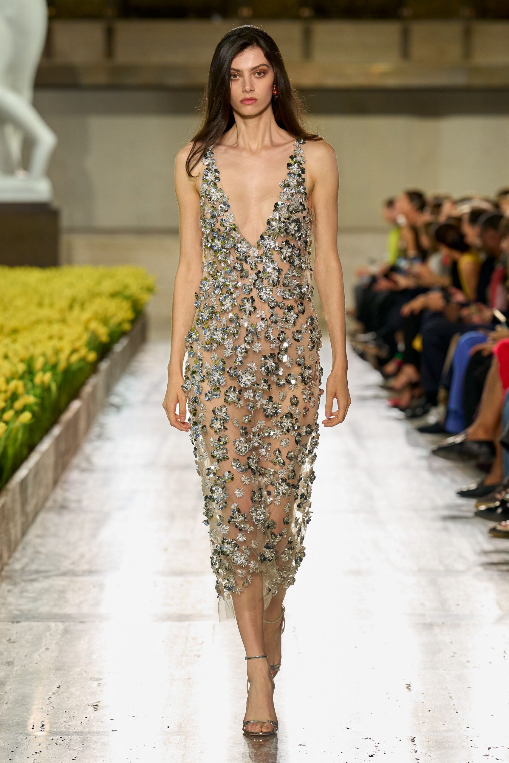 Sheer nude column dress crusted with silver floral paillettes, plunge cut to the waist. Tiny rust-orange stone drops. Bare metallic ankle-strap sandals. Crossing a sculpture court while the fountain has just been turned off.