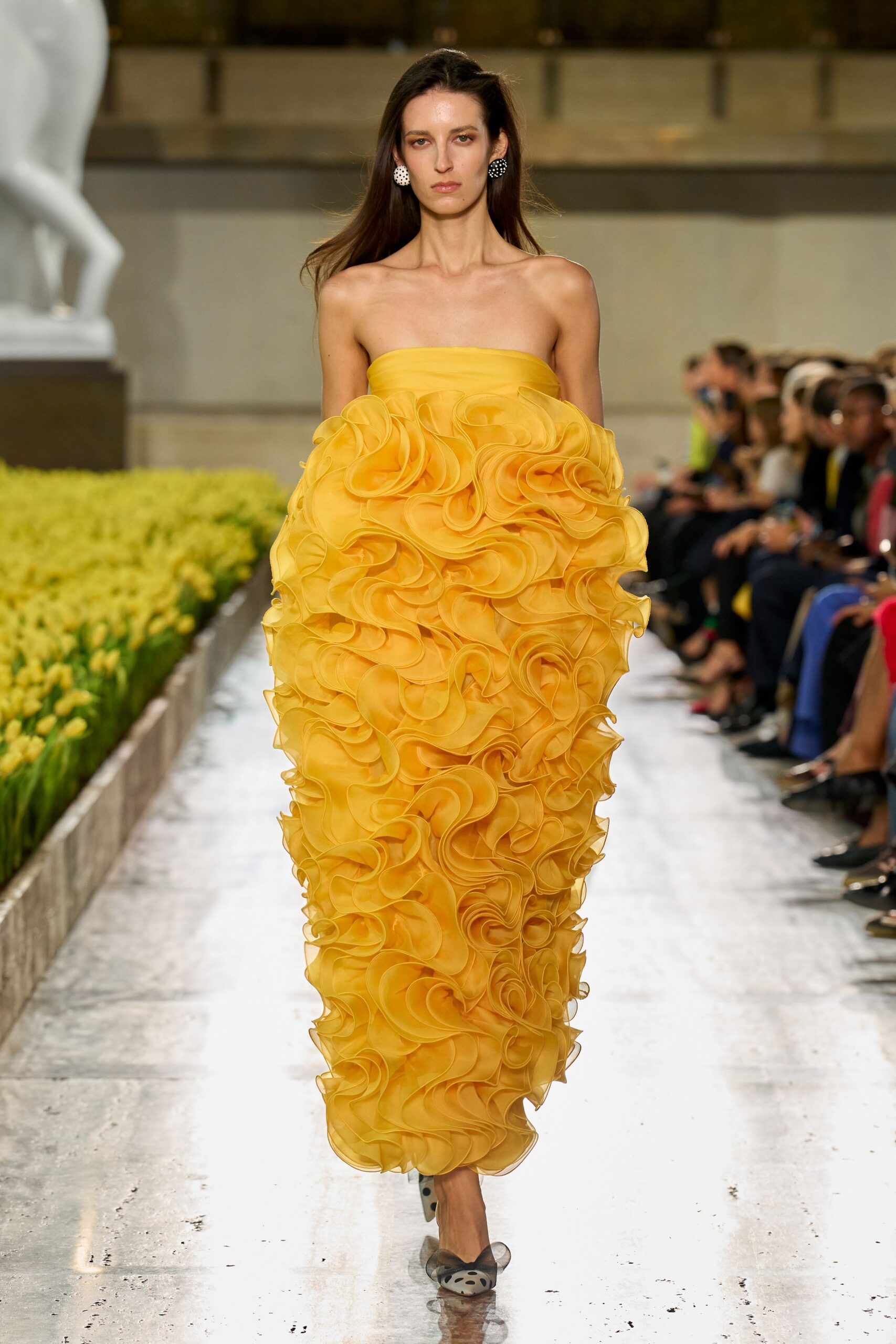 Strapless marigold column gown engulfed in dense organza rosettes and curled ruffles. Round black-and-white polka-dot discs. Sheer mesh pumps with oversized spotted bow fronts. Walking beside a conservatory flower bed at peak tulip week.