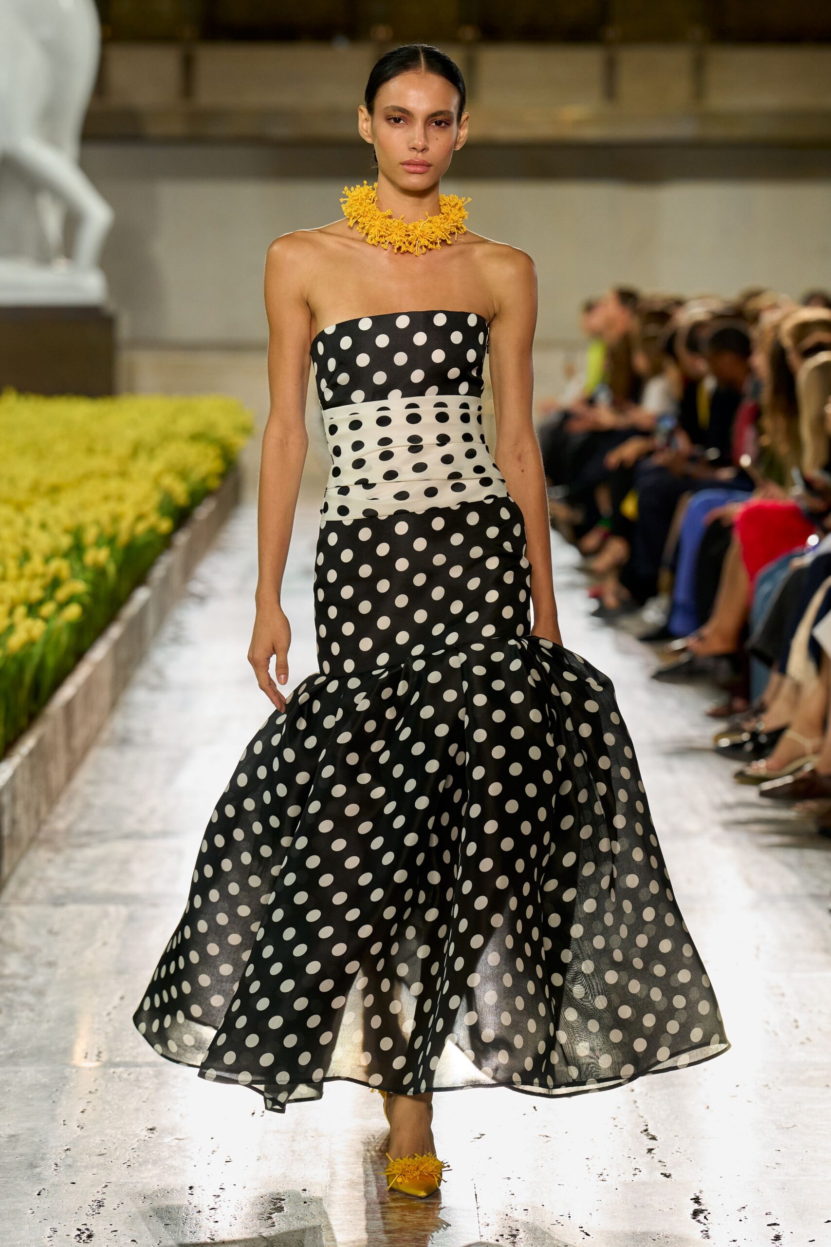 Spiky marigold cluster collar packed tight around the throat. Strapless black-and-white polka-dot gown, ruched white midriff and sheer flounced hem. Satin yellow pumps with shaggy floral tufts at the vamp. Crossing a museum courtyard while the flowerbeds are being replanted.