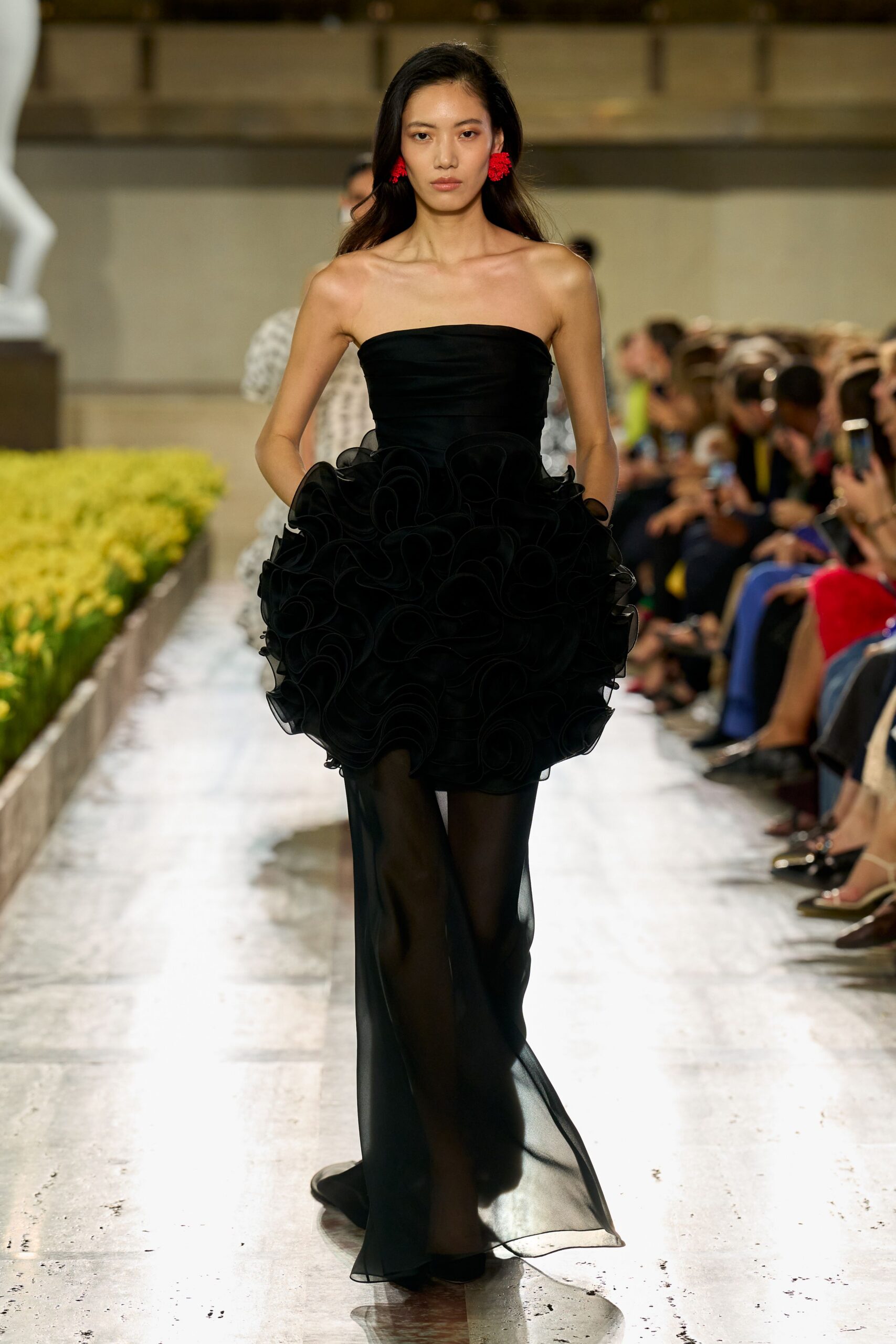 Red tassel cluster earrings brushing the jaw. Black strapless gown, fitted satin bodice swelling into dense organza scrollwork at the hips. Sheer black train falling straight from beneath the bubbled overskirt. Crossing a museum courtyard after dark while the fountain is still running.