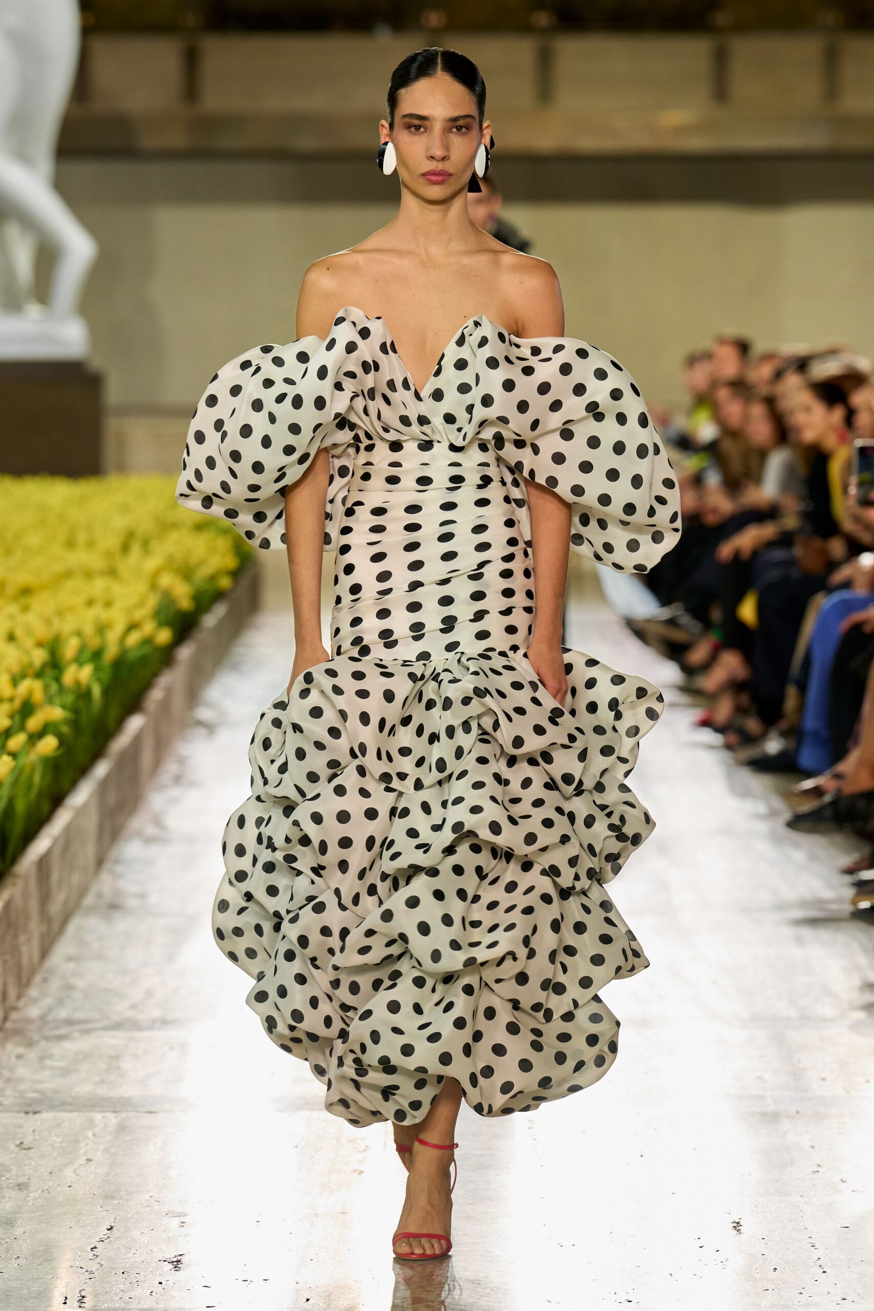 Oversized black-and-white discs clipped close to the lobe. Ivory polka-dot organza gown with off-shoulder balloon swags and a ruched mermaid body collapsing into dense ruffles. Red ankle-strap sandals, barely visible under the hem. Crossing a sculpture court while the tulips are almost blown out.