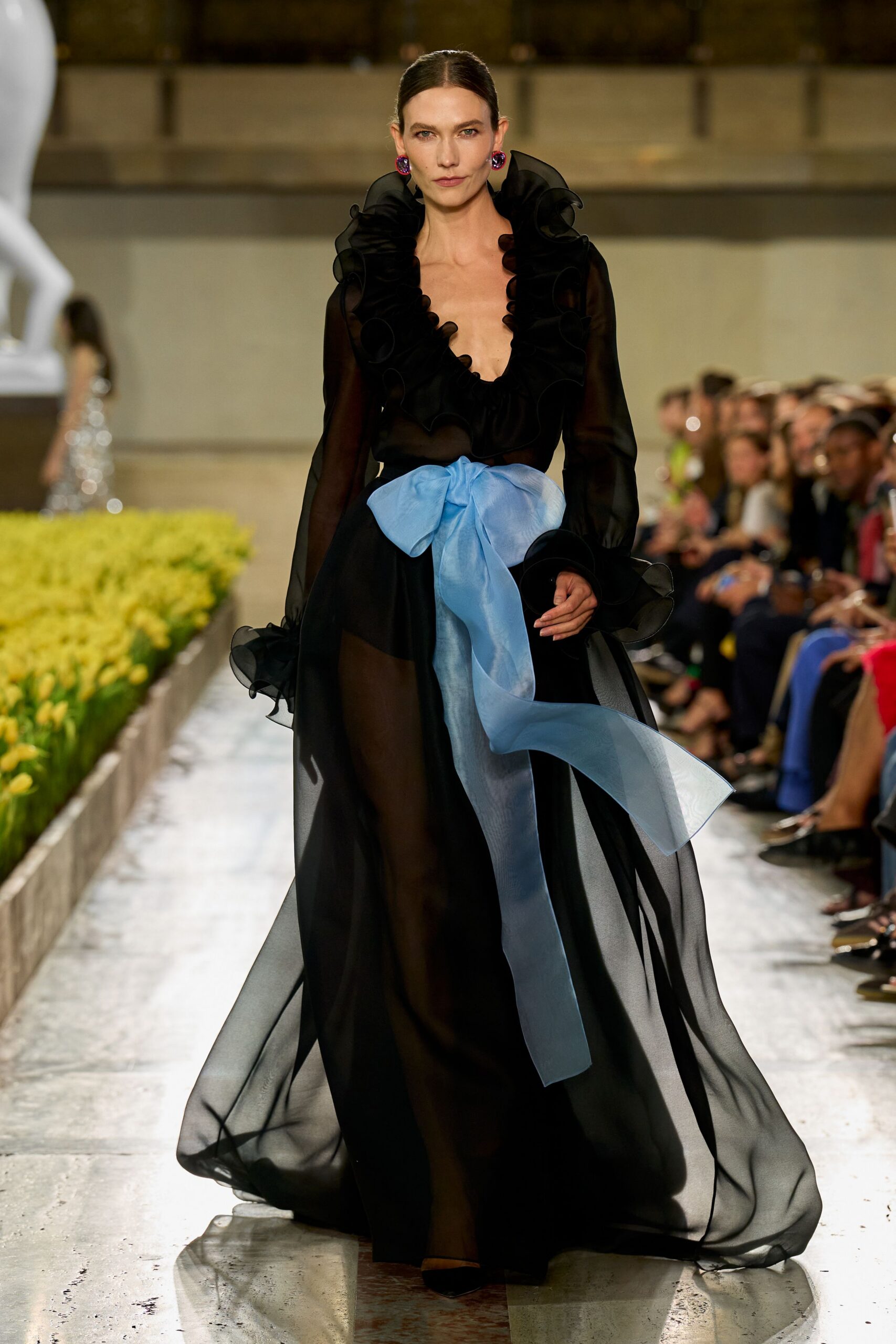 Sheer black chiffon gown with a plunging neck circled in wired ruffles. Powder-blue organza bow knotted at the waist, one tail dragging low. Round violet drop earrings. Black pointed pumps. Crossing a museum courtyard while the string quartet is still tuning.