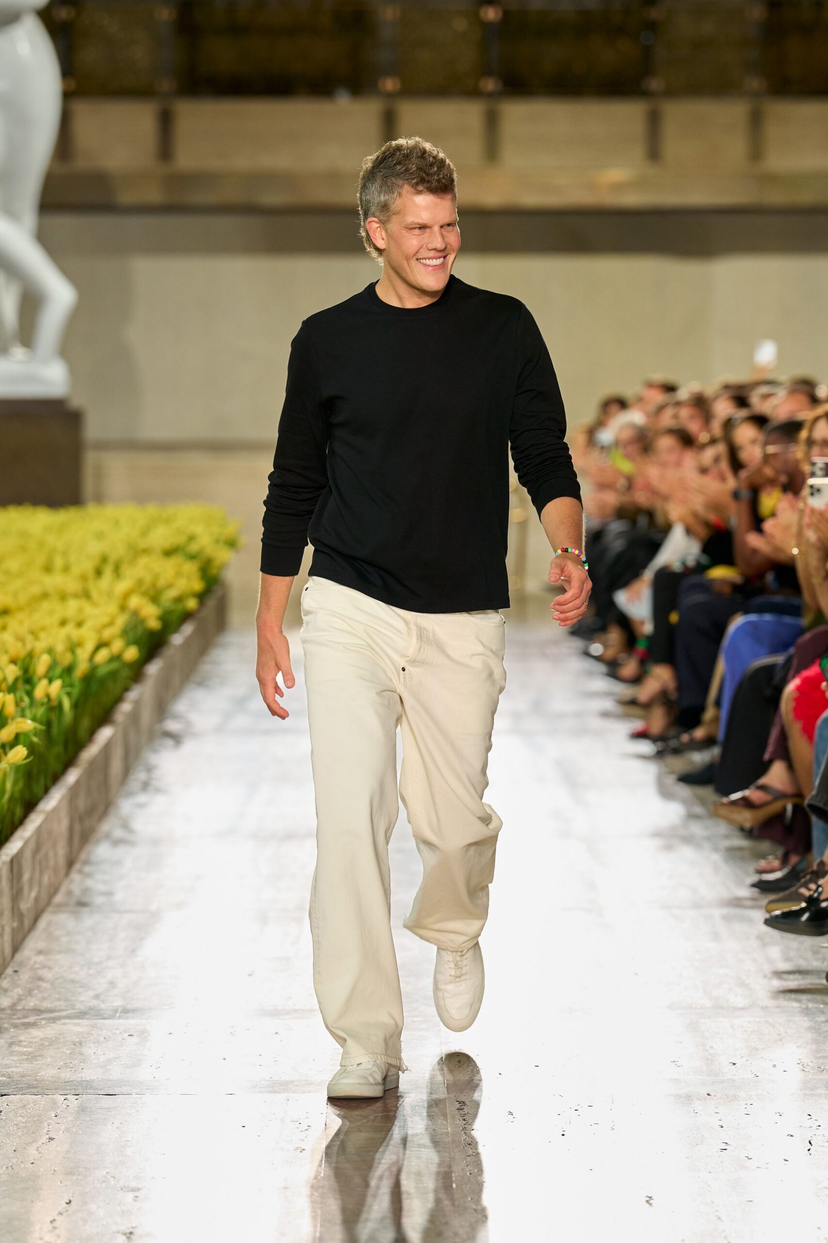 Black fine-gauge crewneck sweater with ribbed cuffs pushed into soft folds. Rainbow bead bracelet at one wrist. Cream trousers in sturdy twill, loose through the leg with a low button fly. White leather sneakers, lightly creased across the vamp. Walking the conservatory aisle after everyone else has gone to lunch.