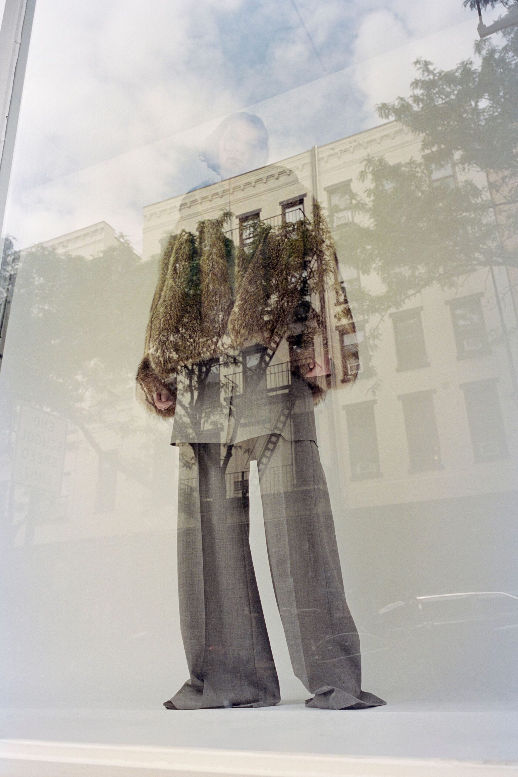 Shaggy straw-brown fur coat hanging to mid-thigh, sleeves swallowing the hands. Grey wool trousers cut extra long, breaking into soft folds over the shoes. Window-shopping on a cold block just before the florist opens.