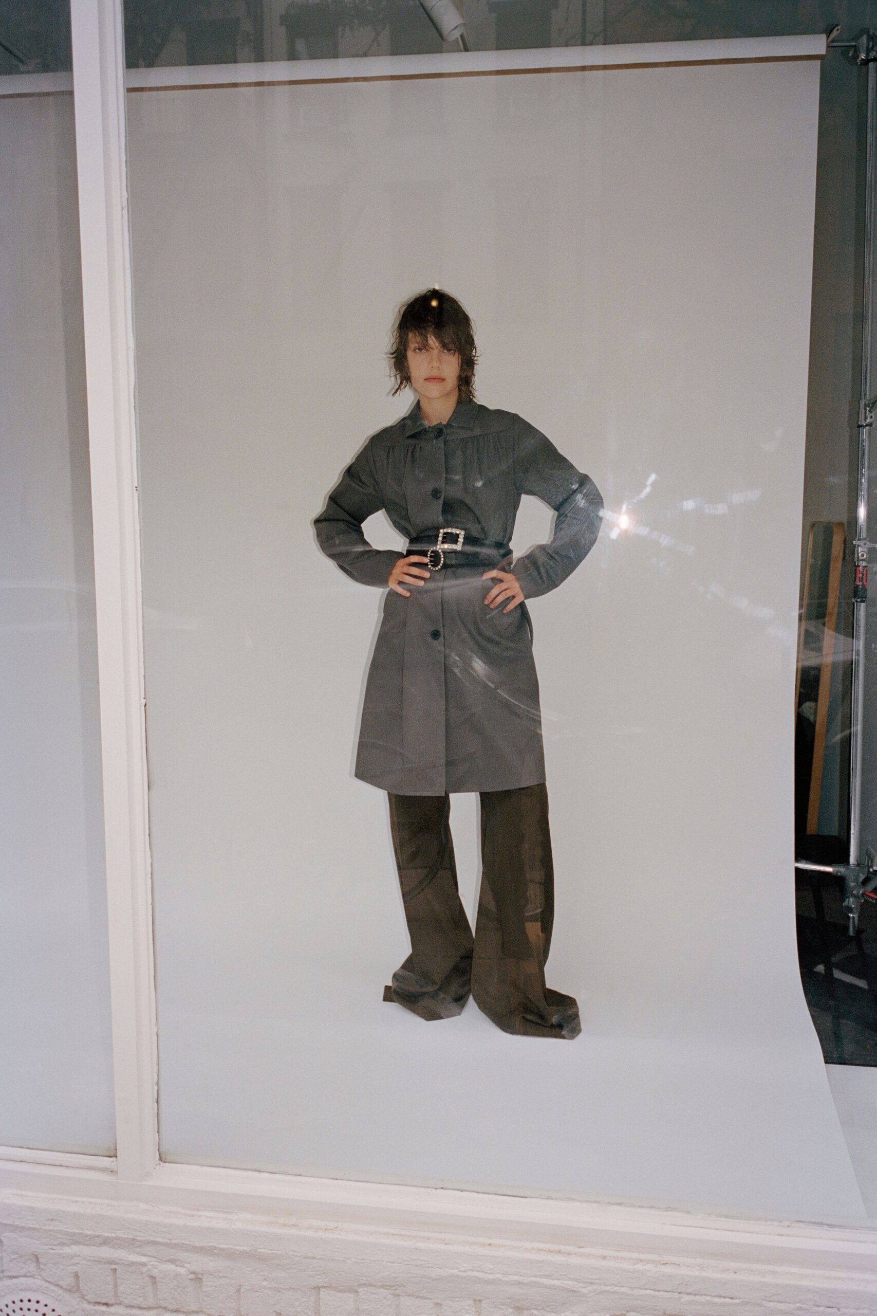 Charcoal coatdress with gathered yoke and oversized black buttons. Wide black belt with rhinestone buckle and oval concho. Dark draped trousers pooling over the feet in soft folds. Afternoon fitting light bouncing off the shopfront glass.