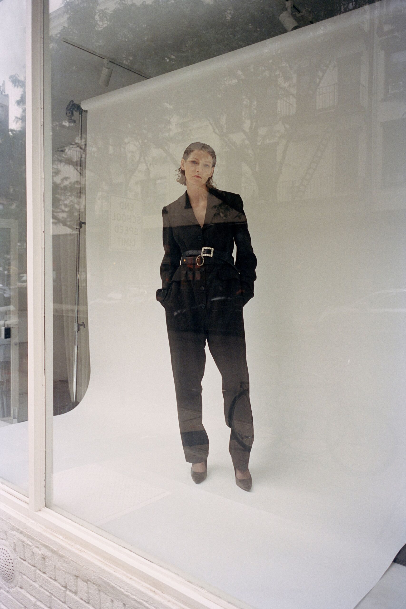Black wool jacket with peaked lapels and a softly flared peplum. Wide black belt set with two rhinestone buckles. Black tapered trousers, slouching slightly above the ankle. Pointed black pumps in matte suede. Window-glass afternoon, trying on your serious voice before the meeting upstairs.