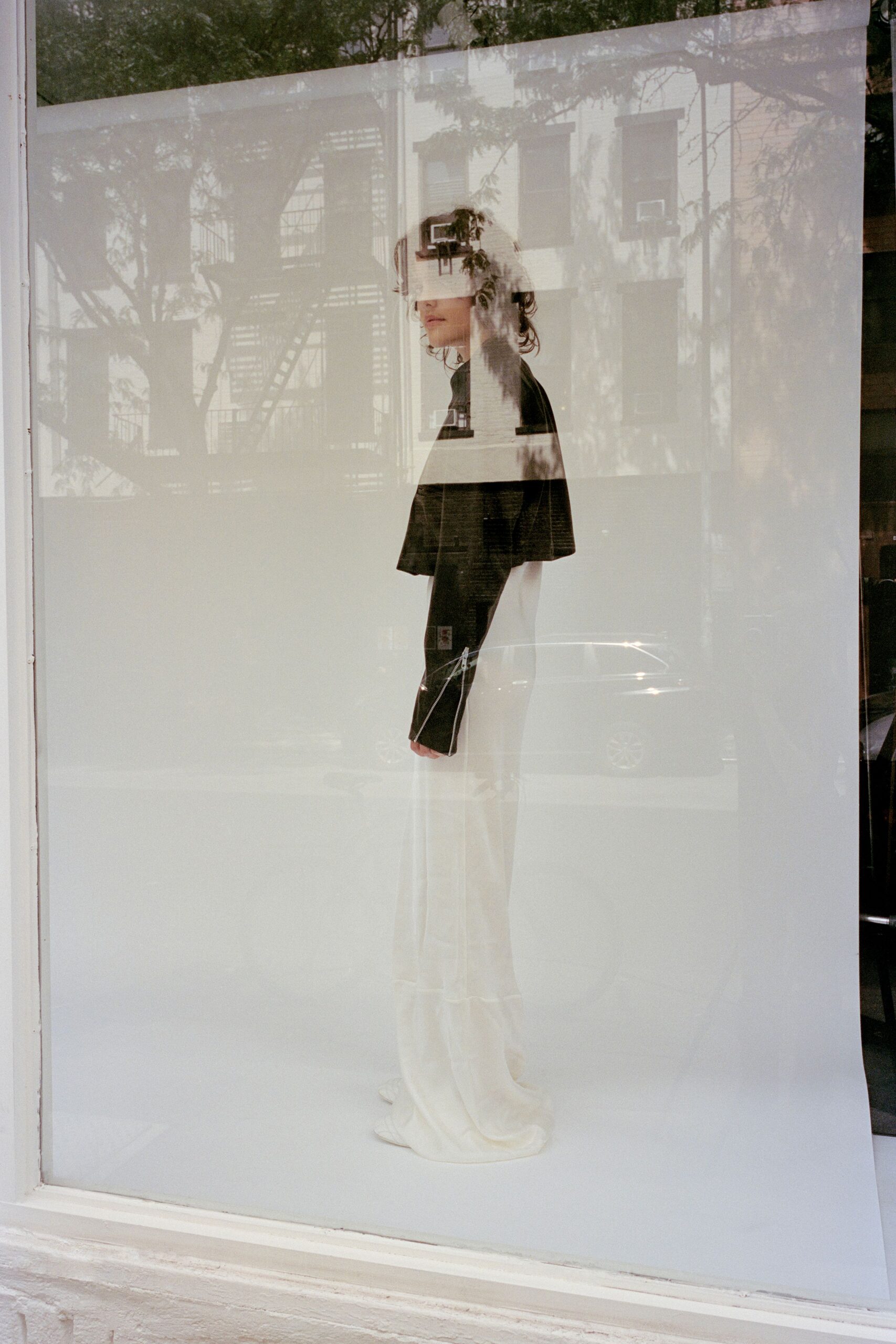 Opaque white visor strapped flat across the eyes. Boxy black jacket with a hard horizontal white band. Cream floor-length trousers breaking into a soft puddle. Pointed white boots peeking from the hem. Window glass at noon, your reflection layered over the opposite building.