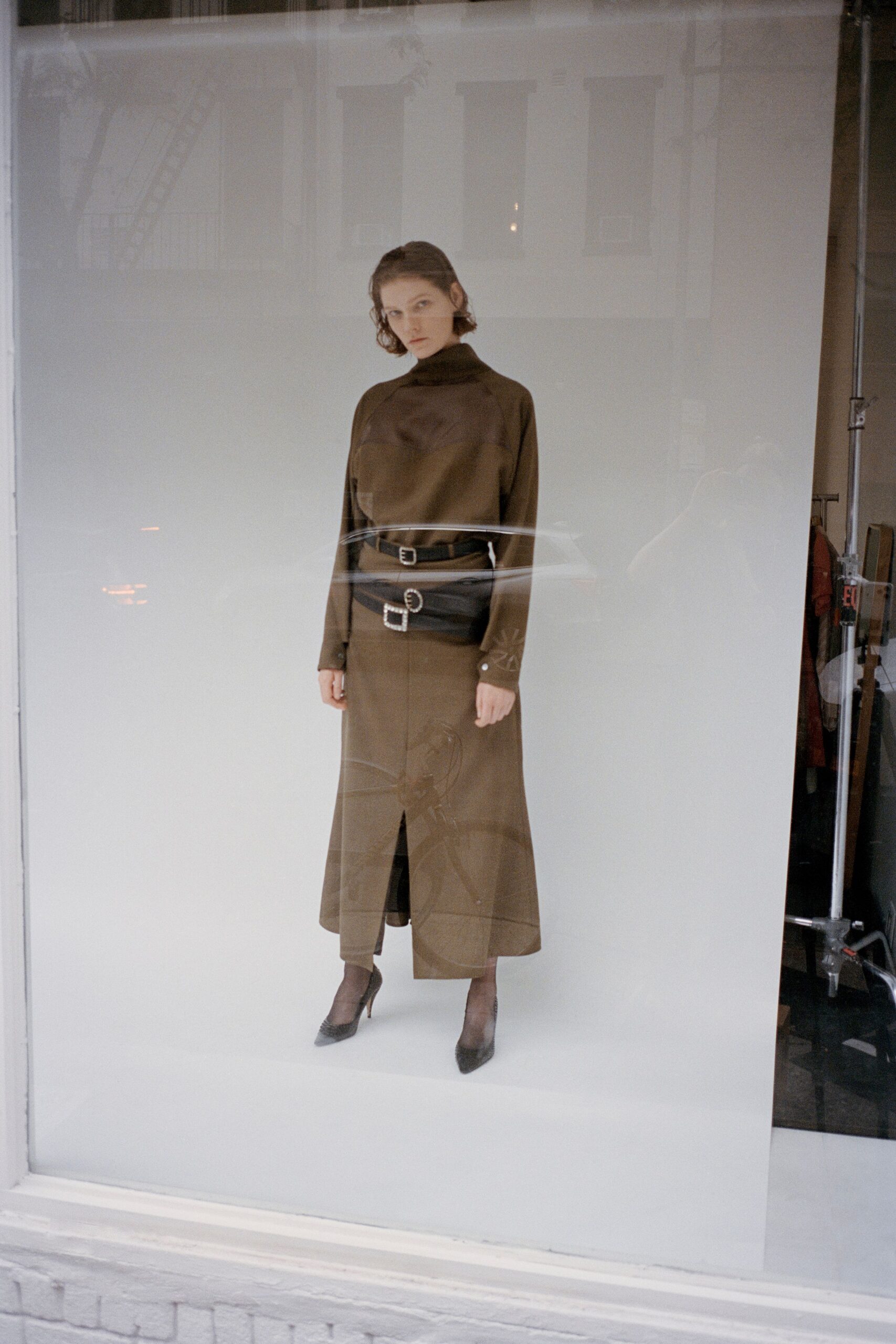 Olive satin mock-neck top with raglan seams and buttoned cuffs. Black leather belt bag cinched twice, two square silver buckles. Matching olive column skirt with a narrow front slit below the knee. Sheer brown stockings. Black mesh pumps with pointed toes. Standing in a shop window after rain, traffic streaking across the glass.