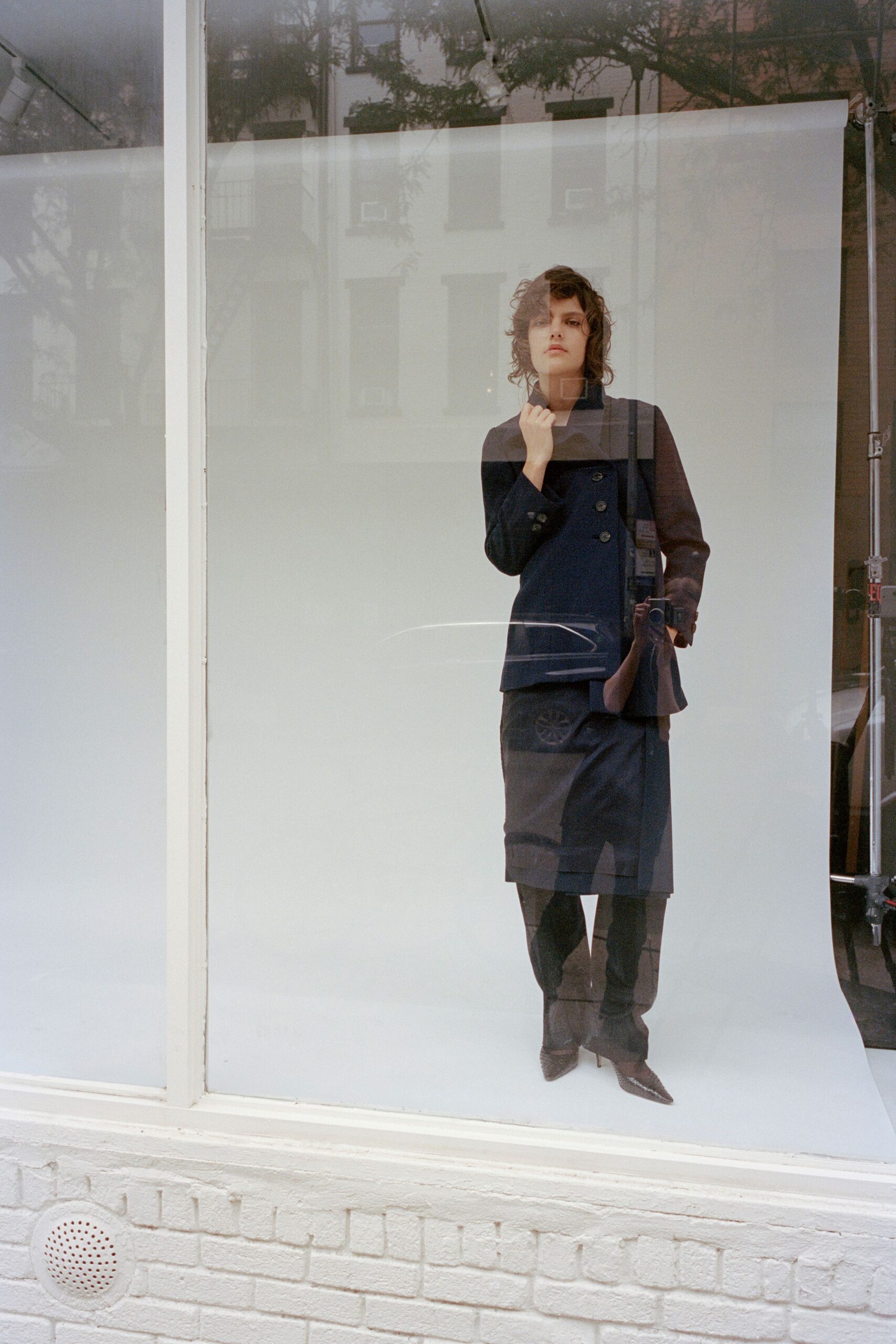 Double-breasted black coat with a high stand collar and flared hem. Straight black knee skirt, lightly creased across the front. Opaque black leggings. Pointed pumps with a fine chain tracing the throat. In a shop window before opening, street trees ghosting across the glass.