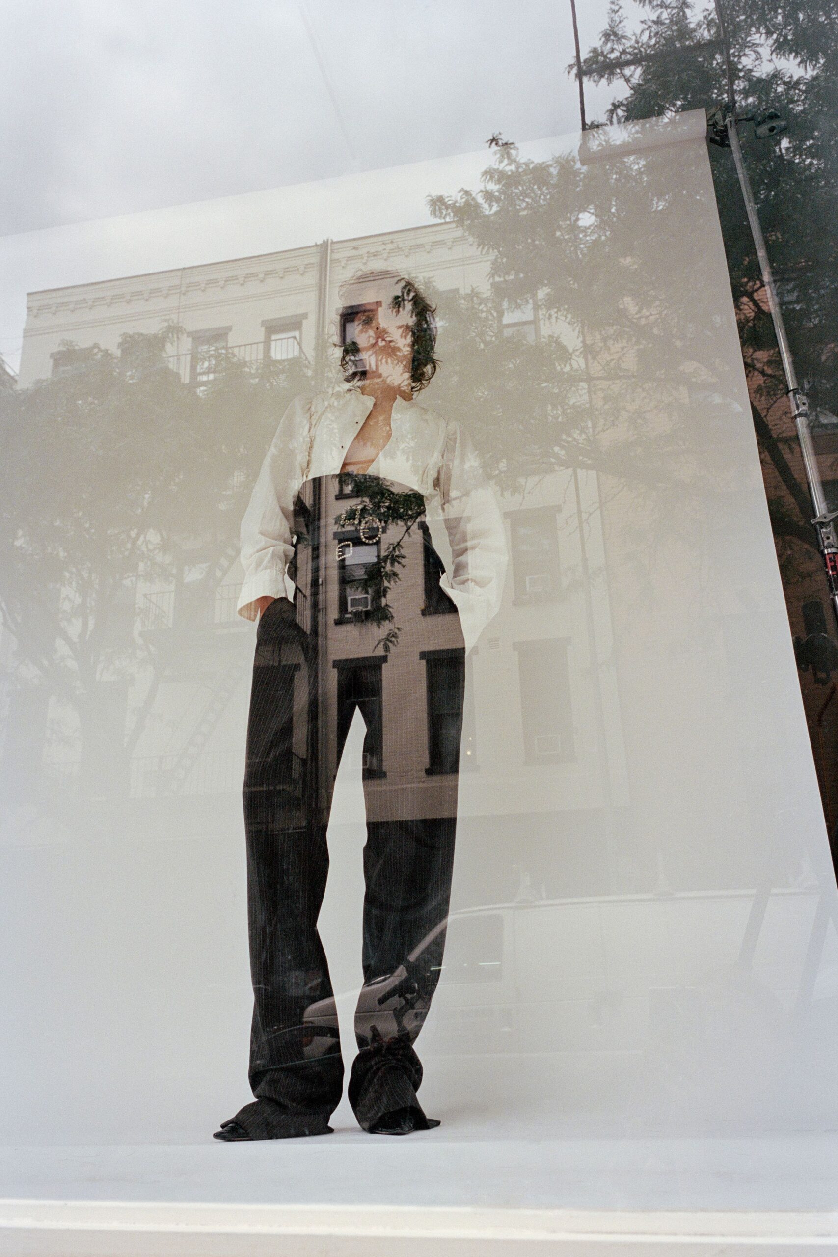 Ivory blouse cut plunging open, sleeves loose and lightly wrinkled. Black corset belt cinched high with two oval buckles. Charcoal pinstriped trousers falling long enough to puddle over the shoes. Black pointed mules, just the toe peeking out. Window glass on a quiet block, late afternoon traffic sliding by.