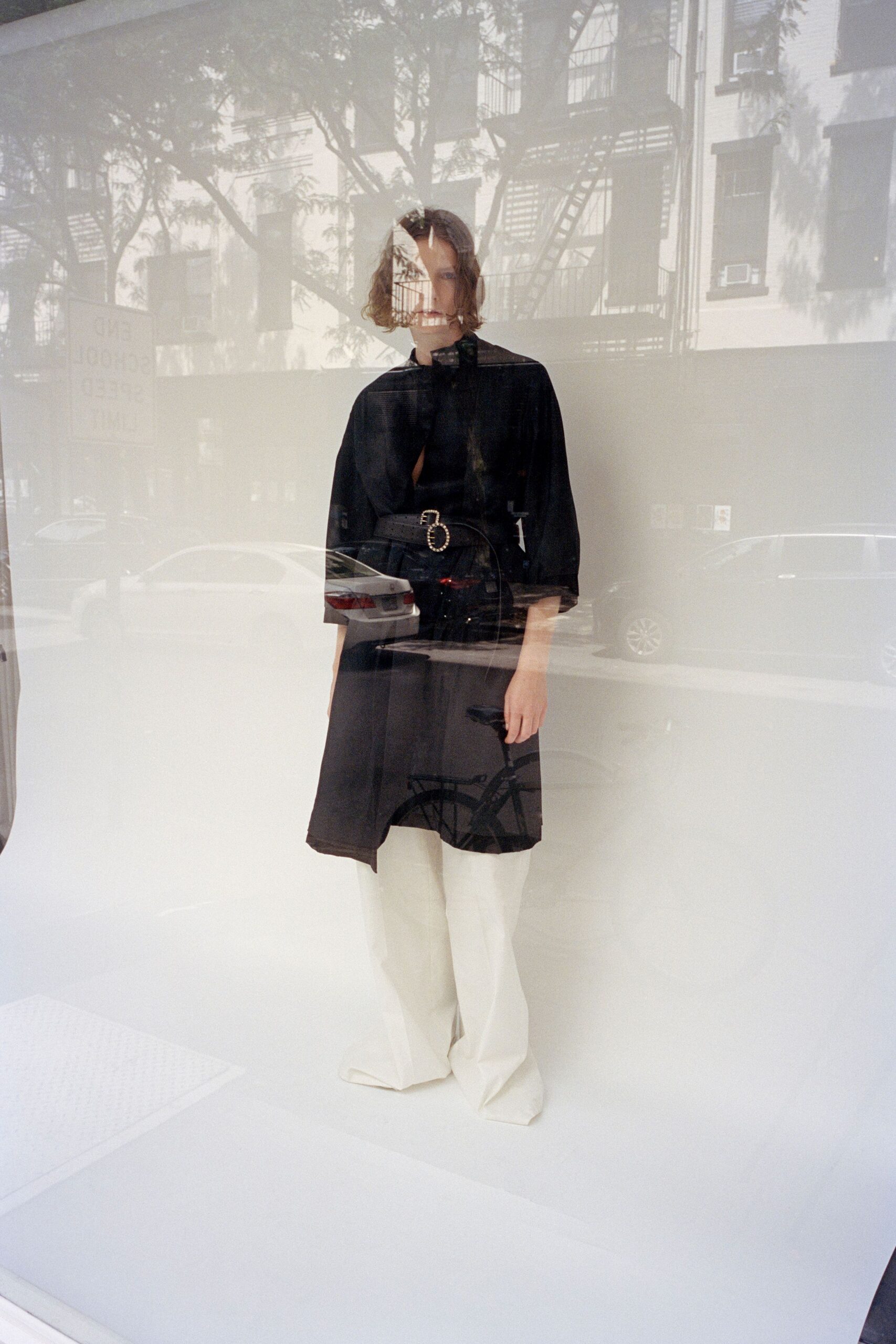 Black matte cape top with a narrow slit opening at the chest. Wide black belt with twin oval buckles set in rhinestones. Cream flared trousers breaking heavily over the shoes. Window glass on a bright city block, traffic sliding over the clothes.