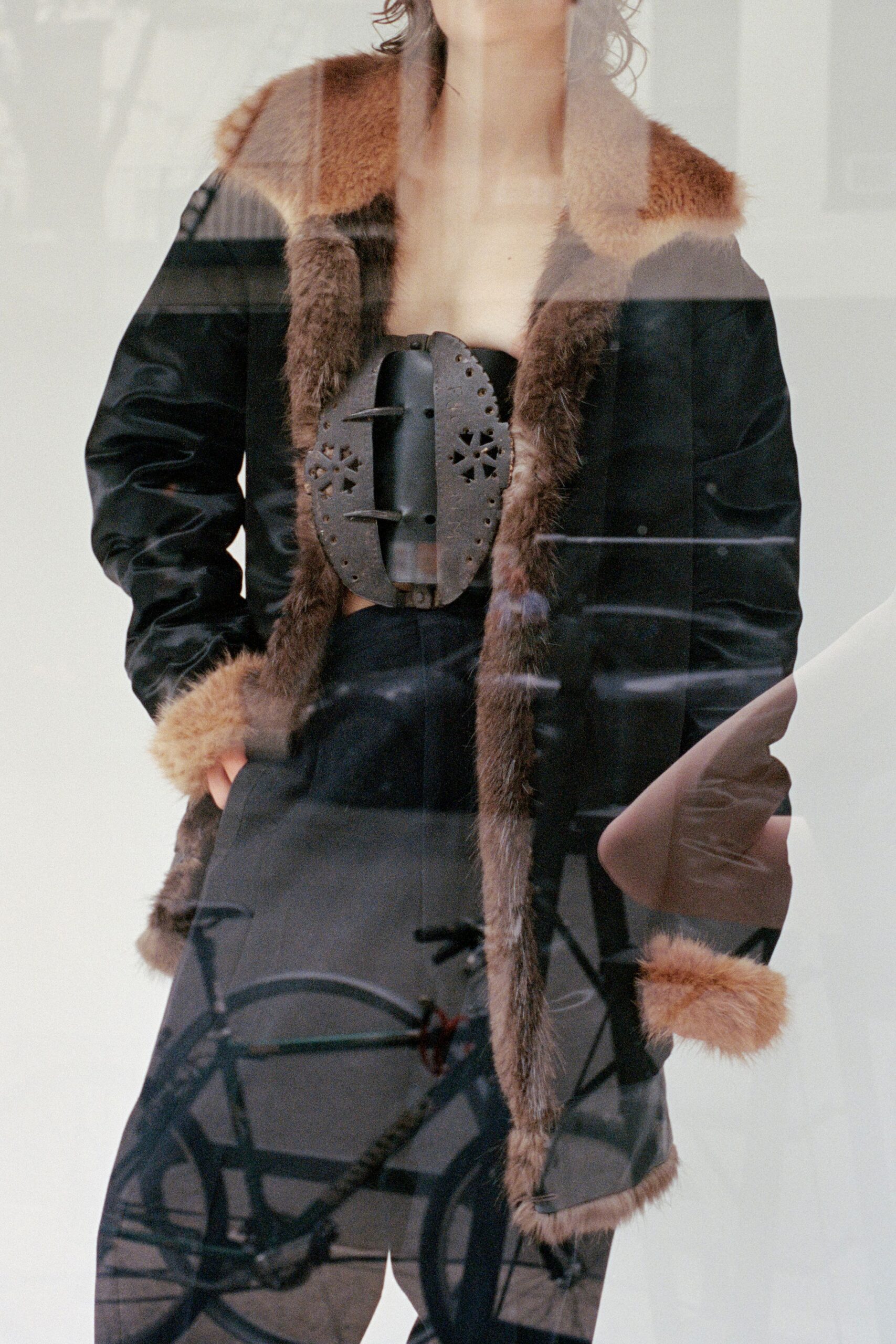 Black leather coat lined and edged in caramel shearling. Strapless black bustier clamped with an oversized pierced metal buckle. Black tailored trousers, crease running clean below the bare midriff. Waiting at a tram stop in first frost, window glass catching the street.