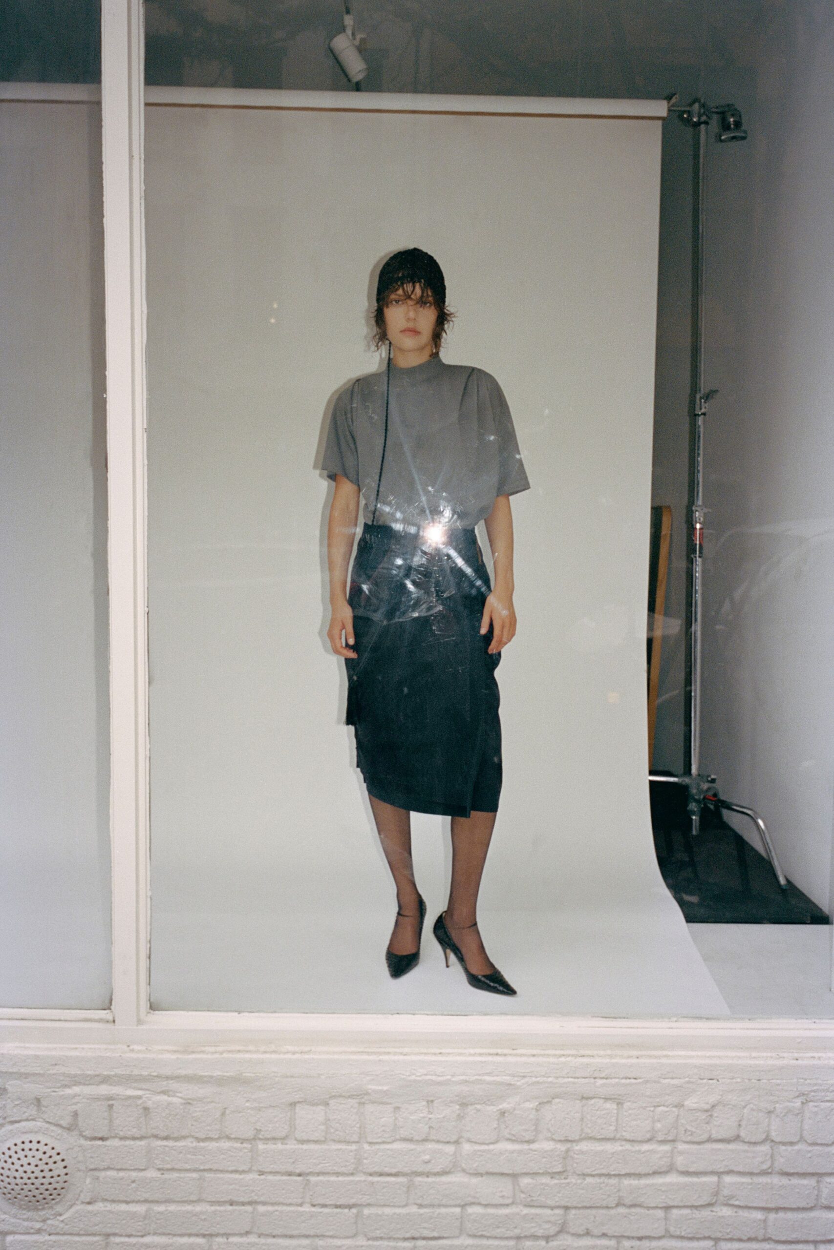 Black crochet beanie with a cord hanging past the shoulder. Dove-grey mock-neck top in glossy jersey, softly bloused at the waist. Black wrap pencil skirt with an offset hem dipping at one side. Sheer black tights. Black pointed pumps with slim ankle straps. Caught in a shop window after rain, deciding whether to keep walking.