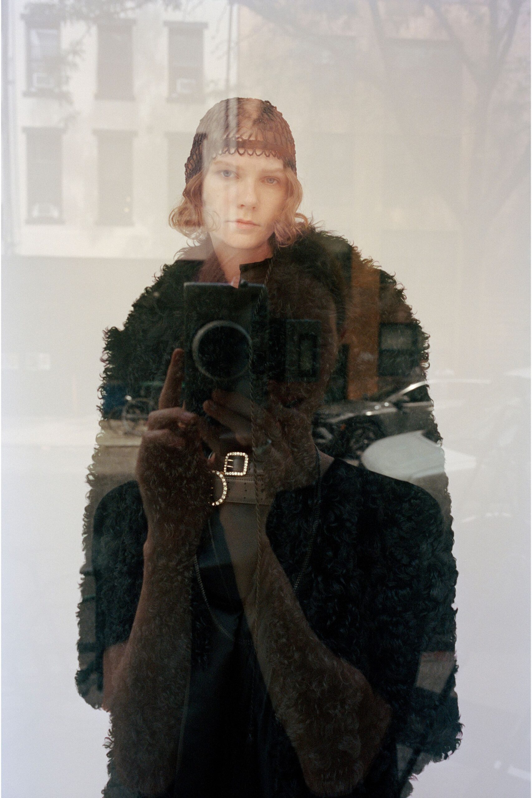 Black crocheted skullcap with a scalloped edge skimming the eyebrows. Inky astrakhan-textured coat, cut boxy through the shoulders. Black underlayer opening to a narrow V at the throat. Wide black belt with twin rhinestone buckles. Window reflection at dusk, scooters parked below the apartment block.