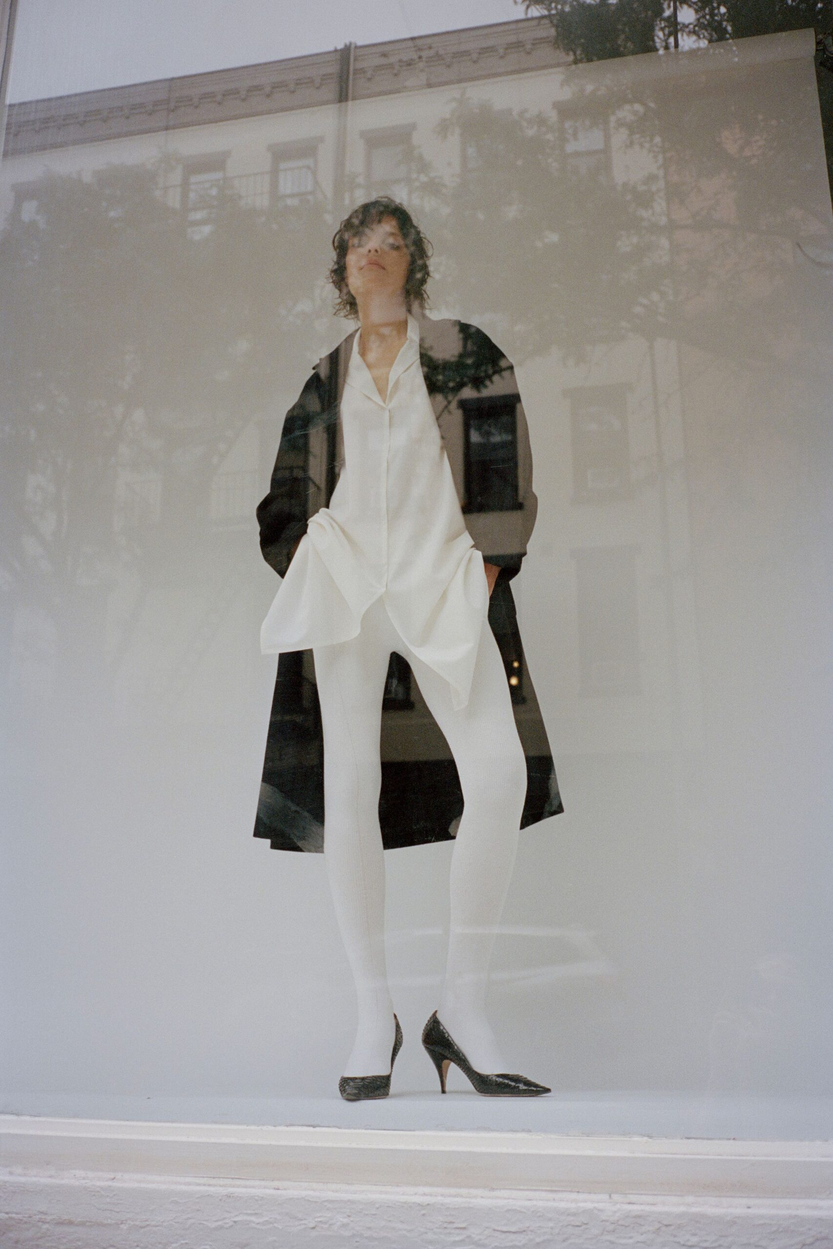 Black knee-length coat worn open, with a clean unbroken front. Ivory fluid blouse with a deep open collar and handkerchief hem. Opaque white ribbed tights, the center seam running straight down. Black pointed pumps in glossy python-textured leather. Window glass full of tree shadows, just before the shop opens.