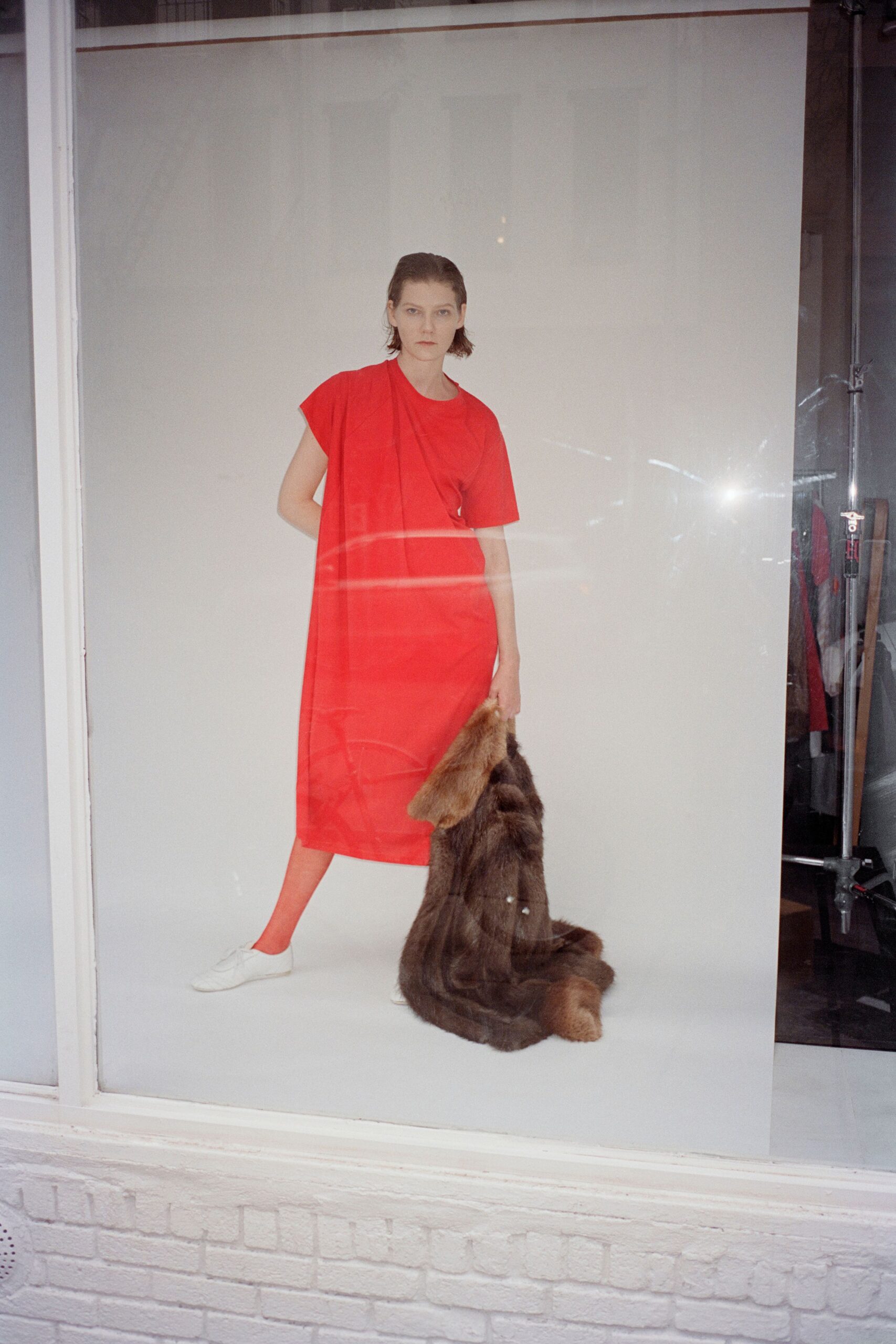Tomato red jersey T-dress with a long panel falling from one shoulder. Matching red opaque tights. White leather lace-ups, plain and low to the ground. Dark brown fur pelt dragged in one hand, tail still hanging. In a shop window at noon, dressed before the heating kicks on.