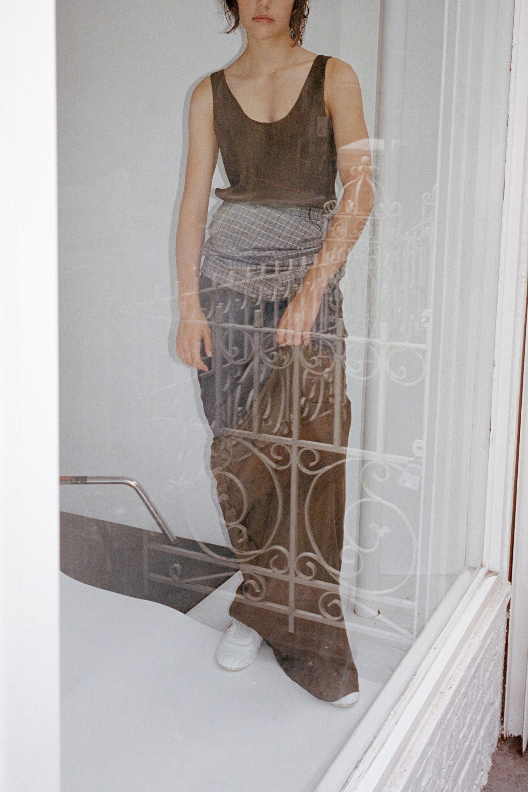 Taupe scoop-neck tank in fine jersey, cut close through the ribs. Grey checked shirting wrapped low and ruched across the hips. Dark brown wide-leg trousers, puddling softly over the shoes. White leather sneakers, rounded toes peeking out. Seen through an apartment window just after the balcony door was opened.