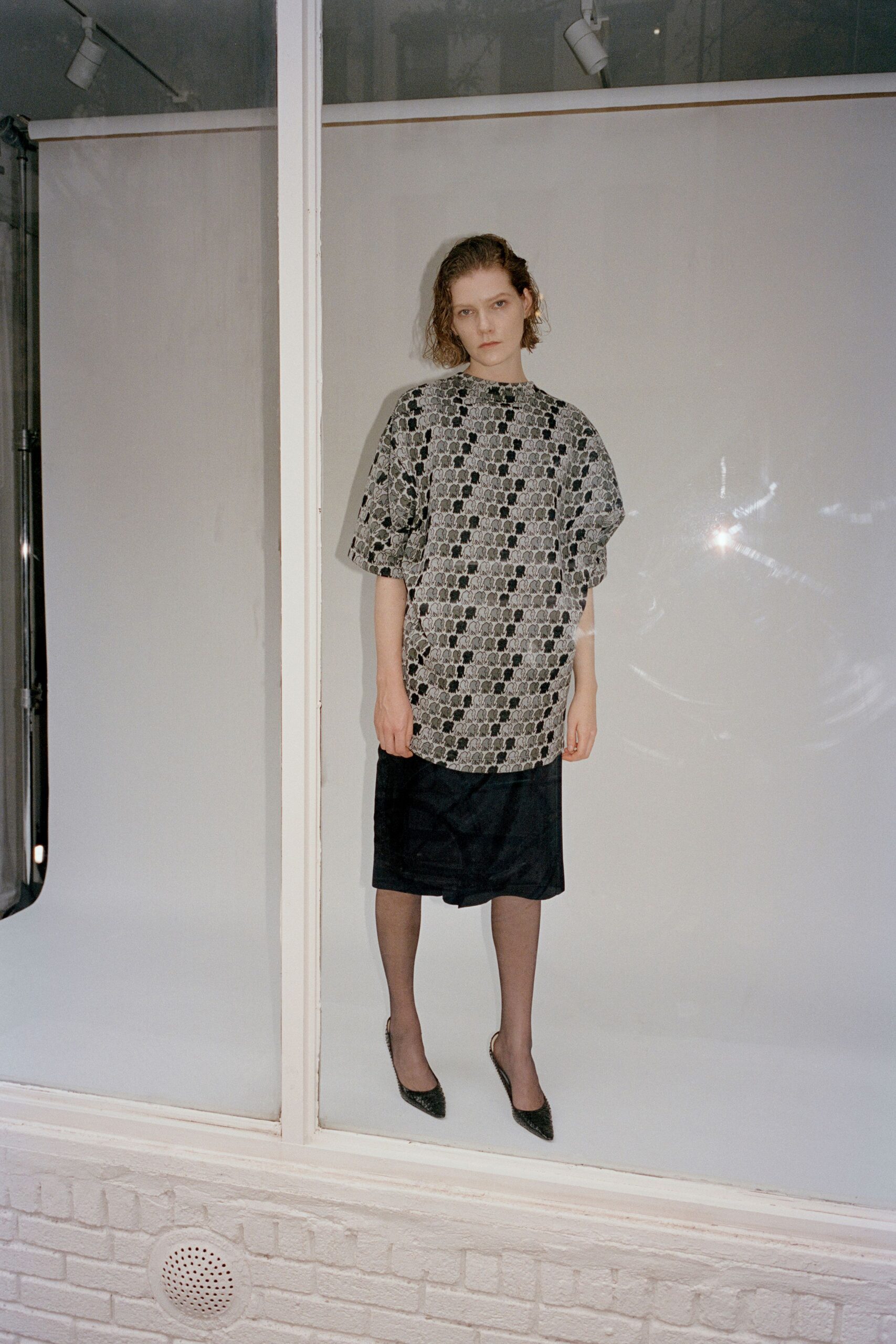 Grey jacquard tunic printed with tiny black elephants, cut to the knee. Black satin wrap skirt, one front corner folding under at the hem. Sheer smoke nylons. Dark woven pumps with narrow pointed toes. Standing in a shop window after closing, the glass still warm from sun.