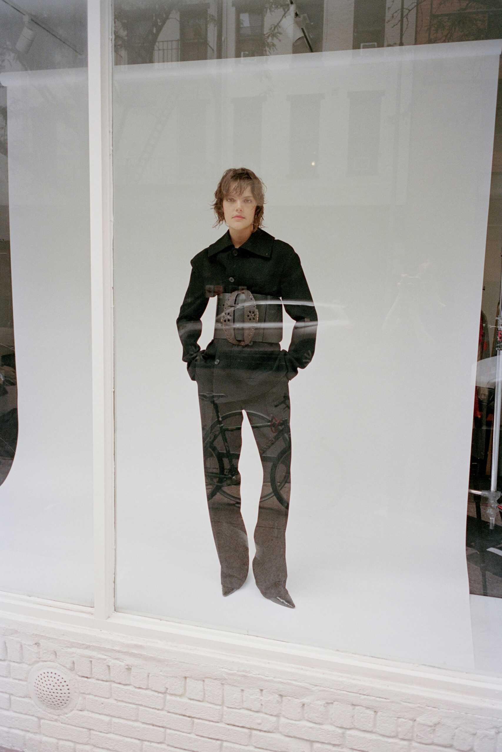 Black cropped jacket with a broad collar and two big buttons. Wide black leather corset belt, punched and laced through an oval front plate. Black straight trousers breaking softly over narrow pointed toes. Standing in a shop window after closing, traffic still streaking past.