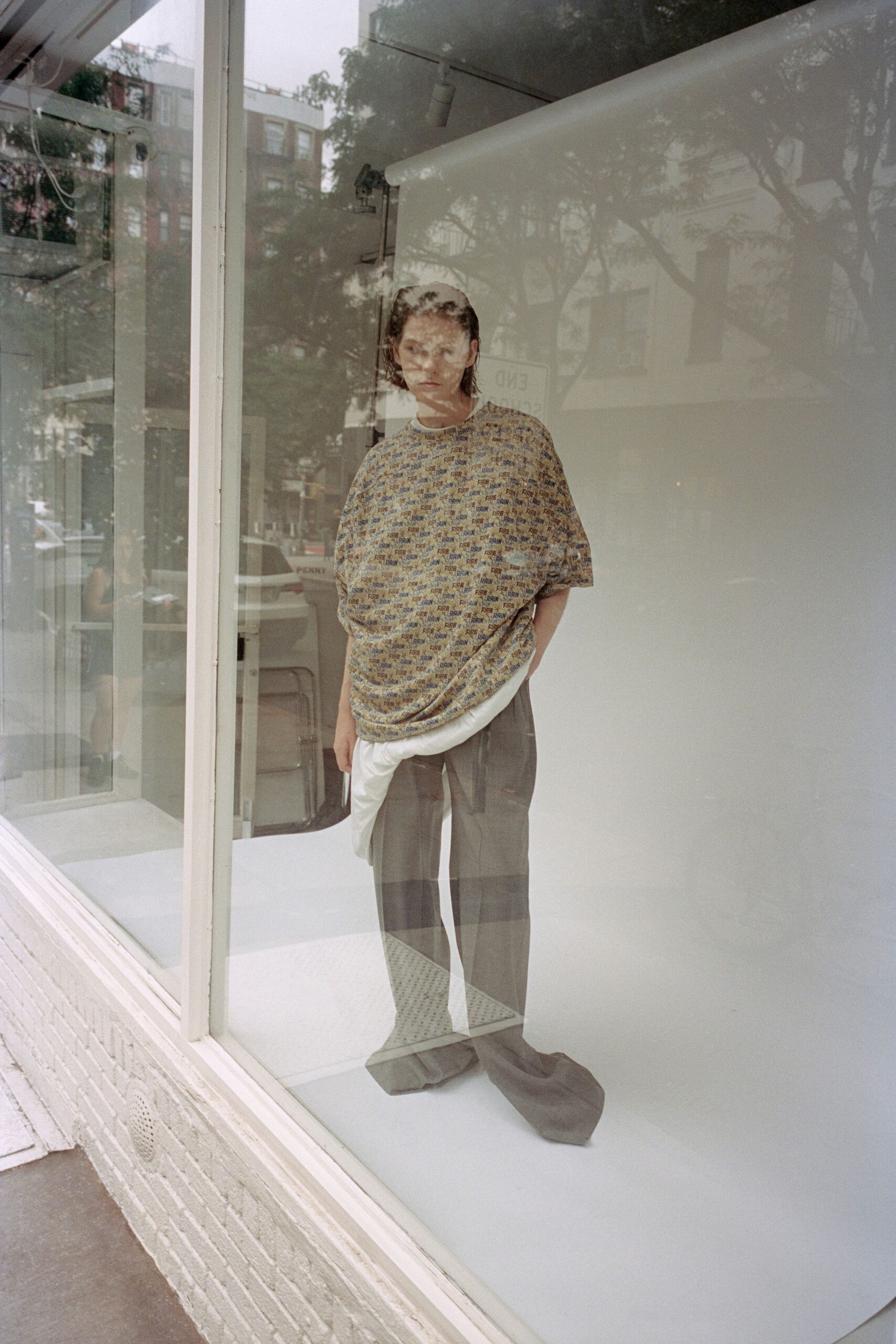 Oversized beige knit tee, tiled with tiny FIRE and RAIN text. White jersey underlayer, scooping below the hem in a thick curve. Ash grey wide-leg trousers with a deep pressed crease and pooled hems. Passing a downtown storefront before opening, your reflection layered with street trees.