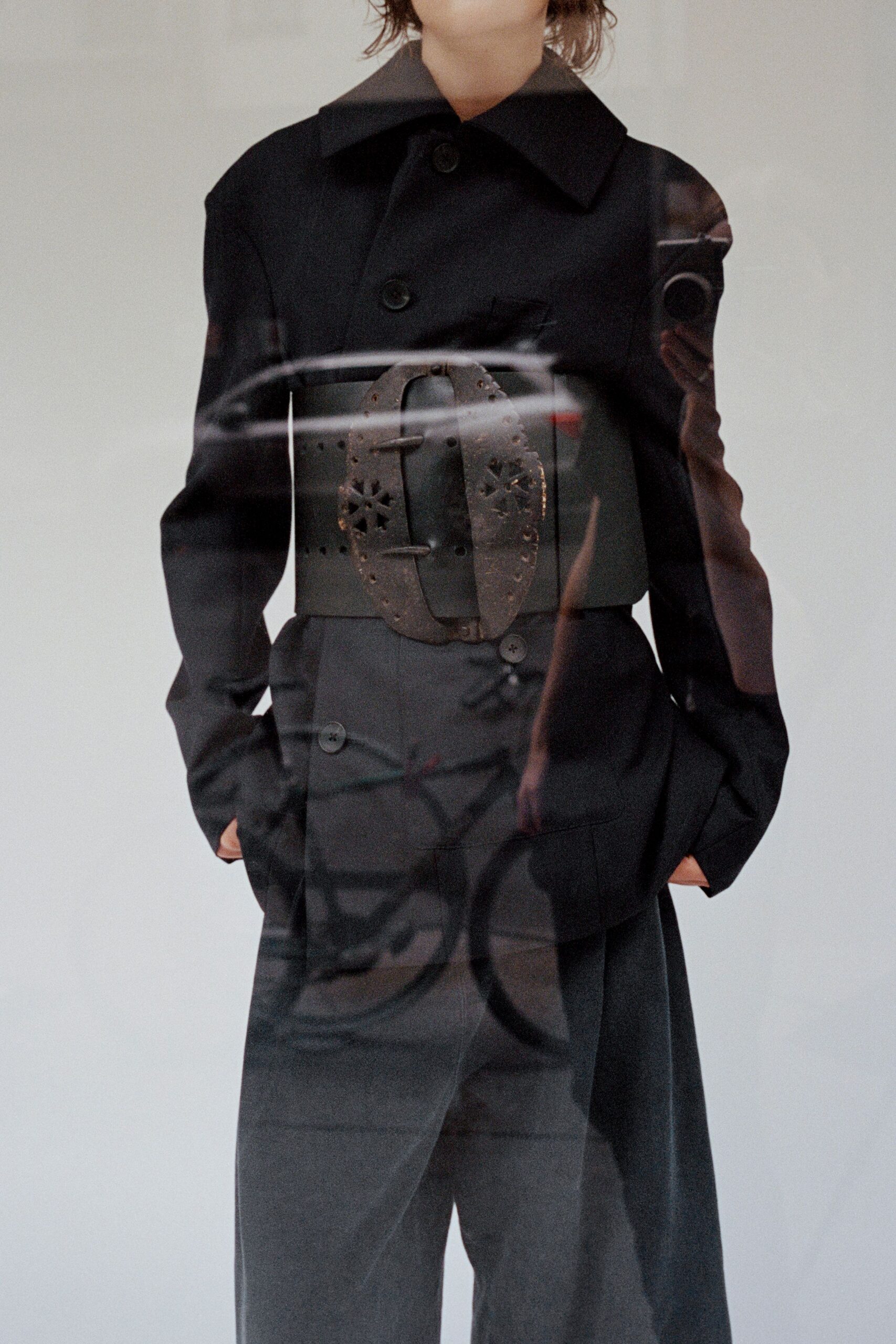 Black tailored coat with an exaggerated spread collar and covered buttons. Wide black waist belt with a pierced metal buckle plate. Dark straight trousers with a double-button tab sitting off center. Crossing the avenue after rain, storefront glass still full of traffic reflections.