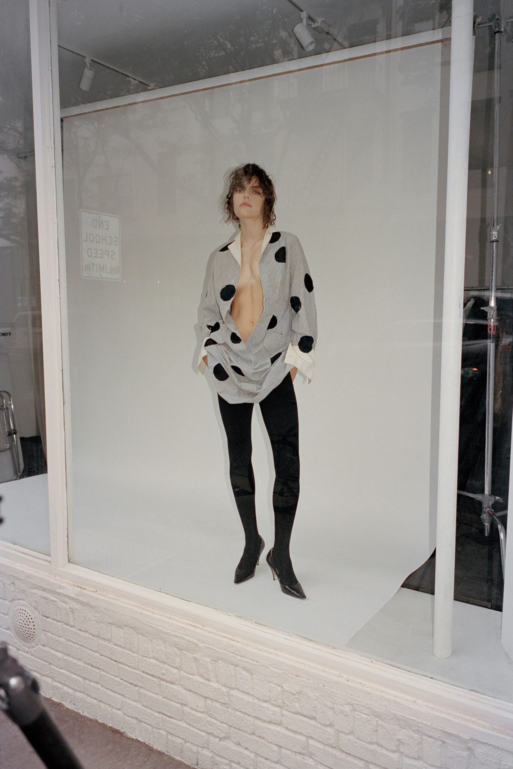 Grey pinstriped blouse scattered with fuzzy black polka dots, plunging to the navel. Opaque black stirrup leggings, glossy at the stretched knees. Pointed black boots in mesh-textured leather with thin stiletto heels. Standing in a shop window before opening, street still wet from drizzle.