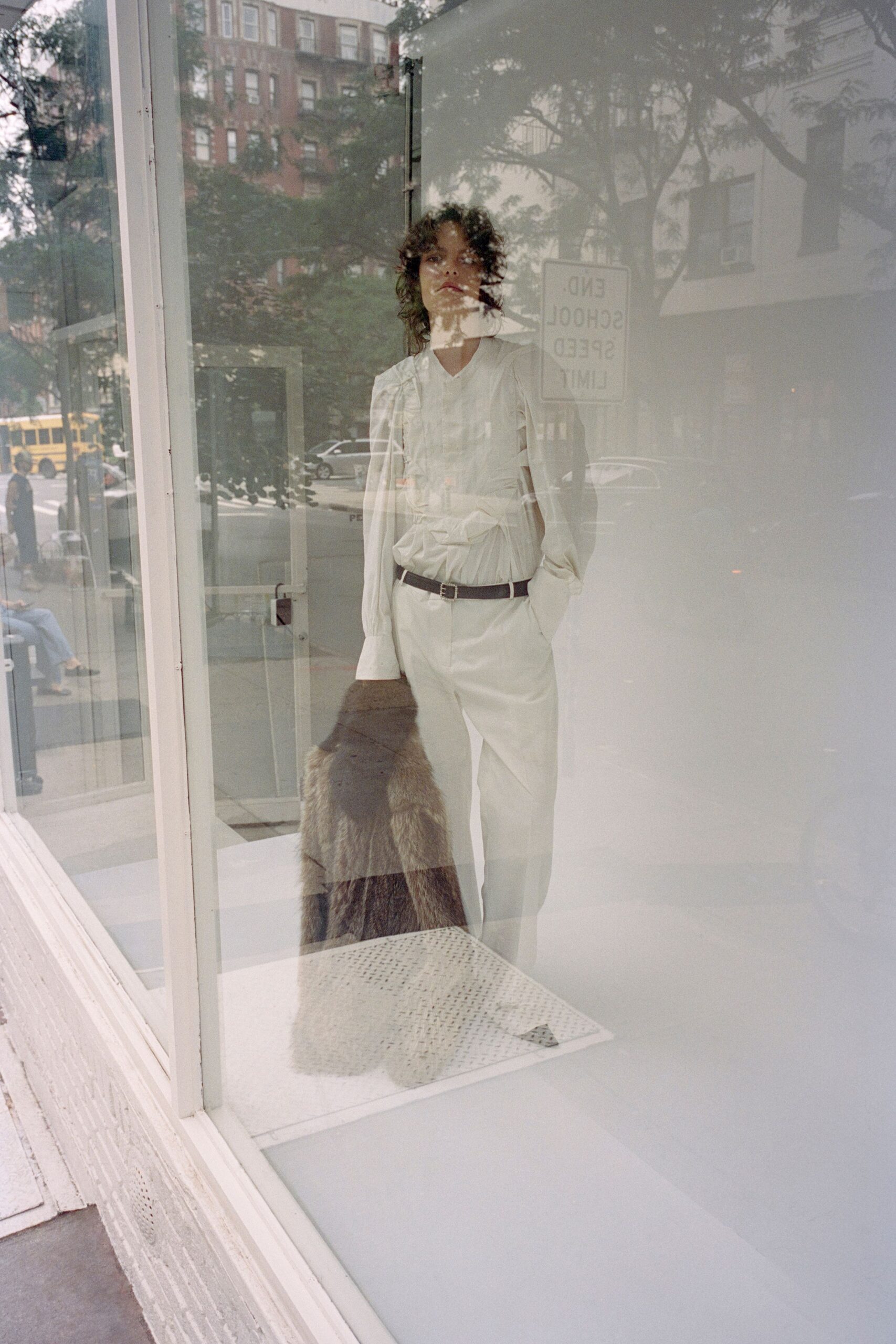 White blouse with gathered seams radiating into a pinched waist. Narrow dark leather belt with a small square buckle. Cream straight trousers, pressed flat and breaking softly at the shoe. Long brown fur stole carried low, striped darker through the pile. Pausing at a school-zone storefront before the street fully wakes.