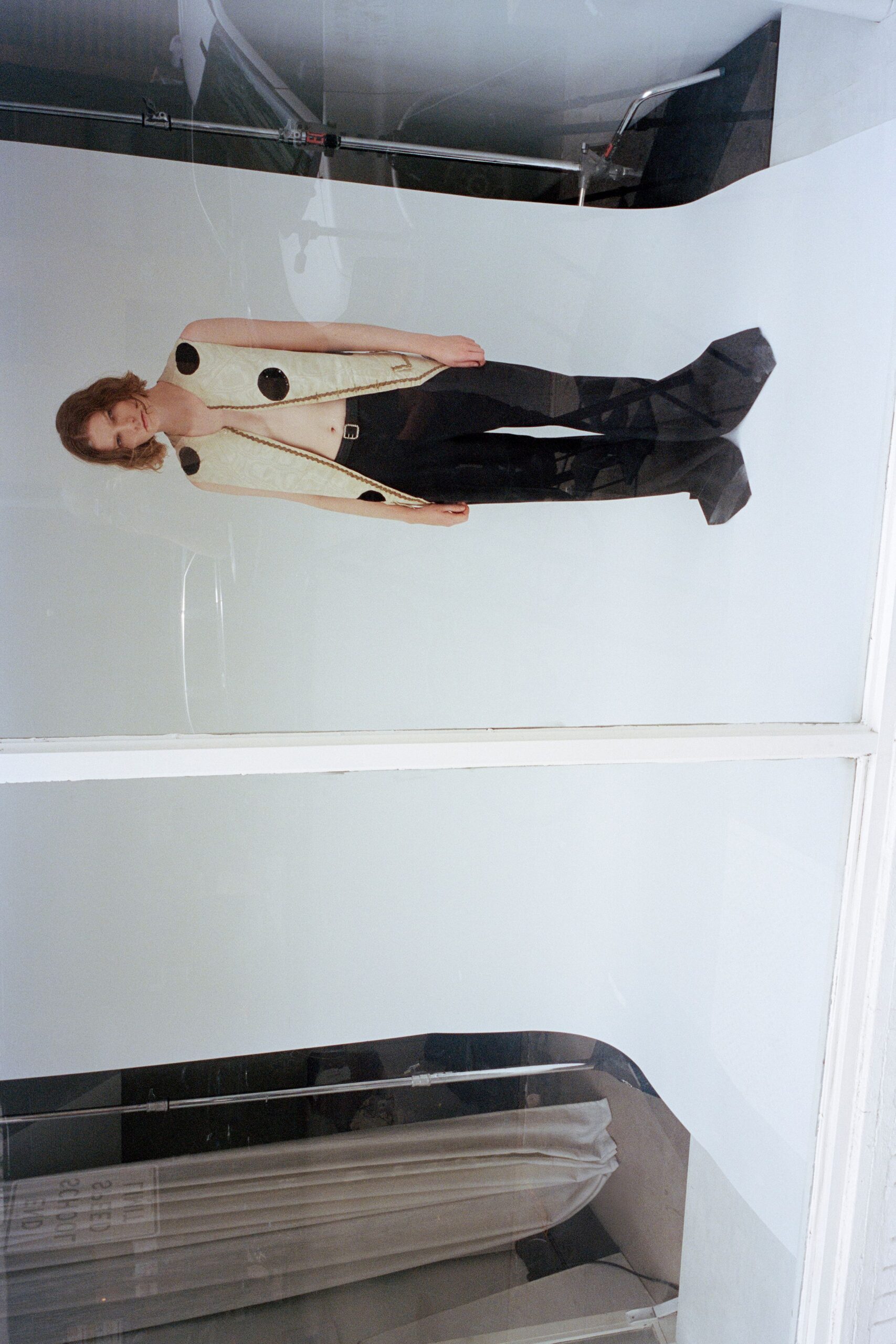 Long cream quilted vest with black circle appliques and whipstitched edges. Narrow black belt with a small square silver buckle. Black sheer trousers with angular faceted overlays flaring over the feet. Walking through a design fair where every chair looks impossible to sit on.