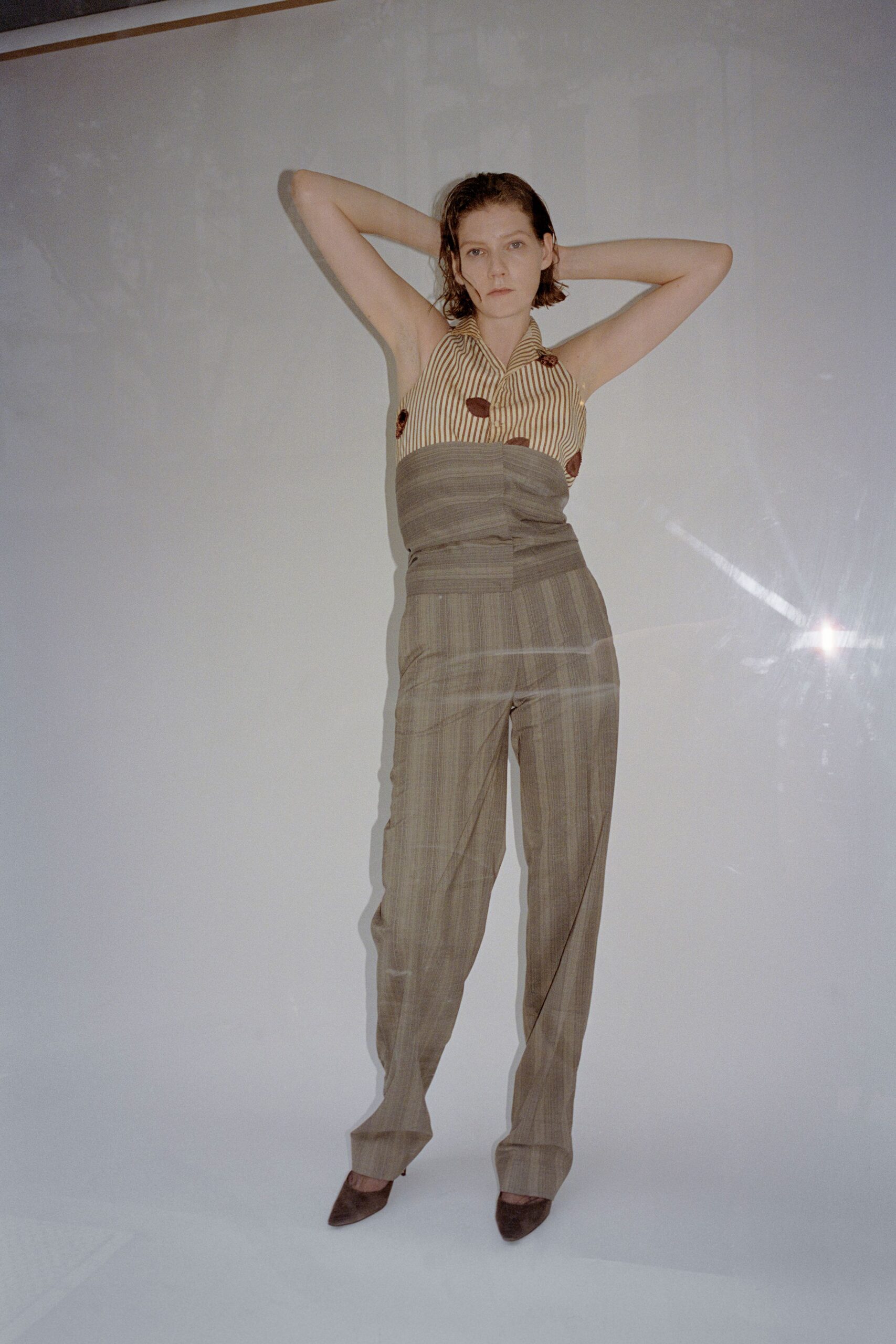 Cream and brown pinstriped halter with scattered velvet spots and a broad lapel. Taupe suiting cloth wrapped high, horizontal seams cinching the waist. Matching taupe trousers with faint chalk stripes and a soft break. Brown suede pointed pumps. Waiting for room service in a hotel still smelling faintly of fresh paint.