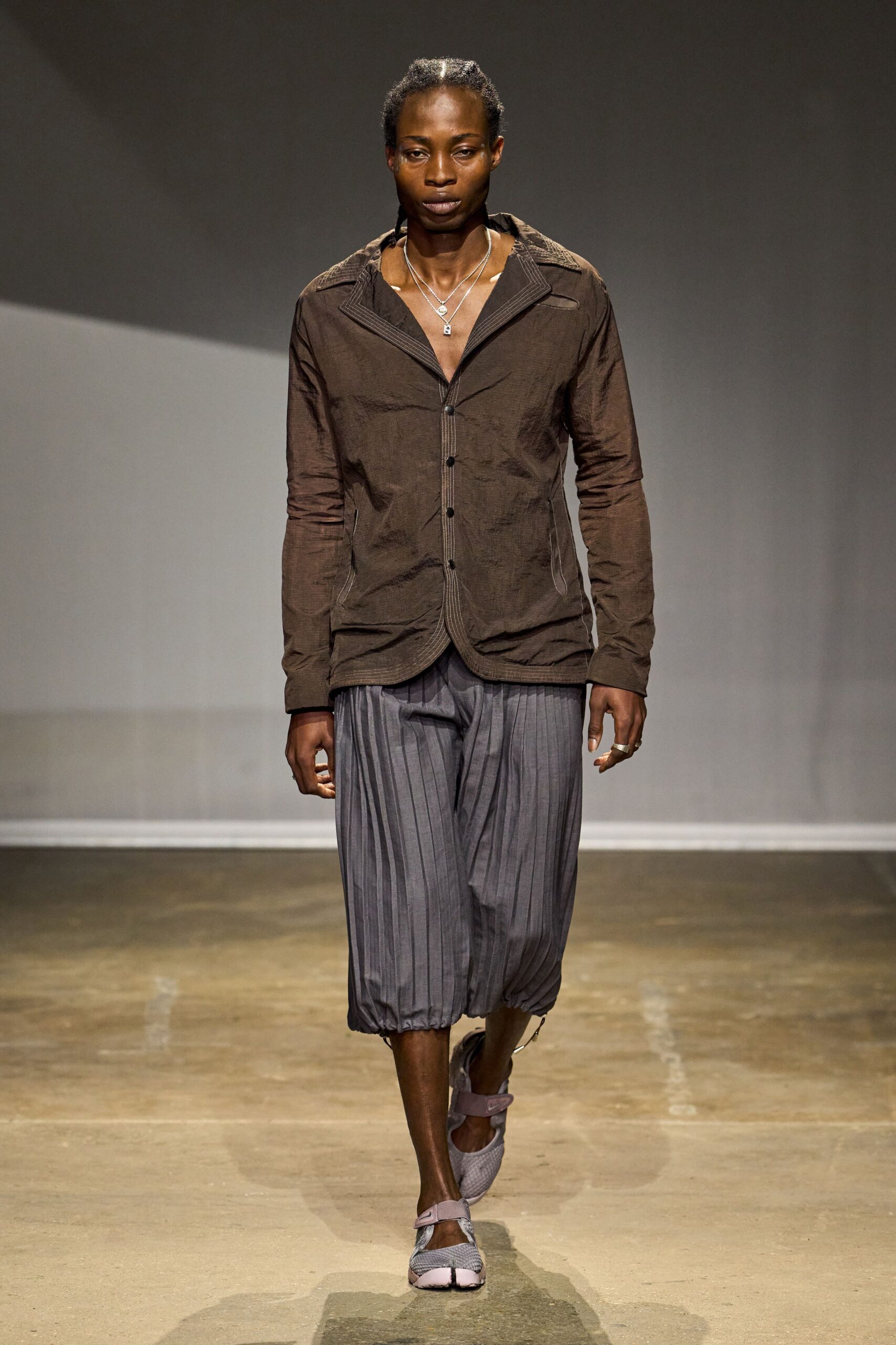 Crinkled espresso shirt with contrast topstitching and a deep open notch collar. Layered silver chains with a tiny padlock pendant. Steel pinstriped culottes, softly pleated and cinched tight at the hem. Split-toe mesh flats with lilac strap closures. Crossing the station concourse after rain, one platform left to check.