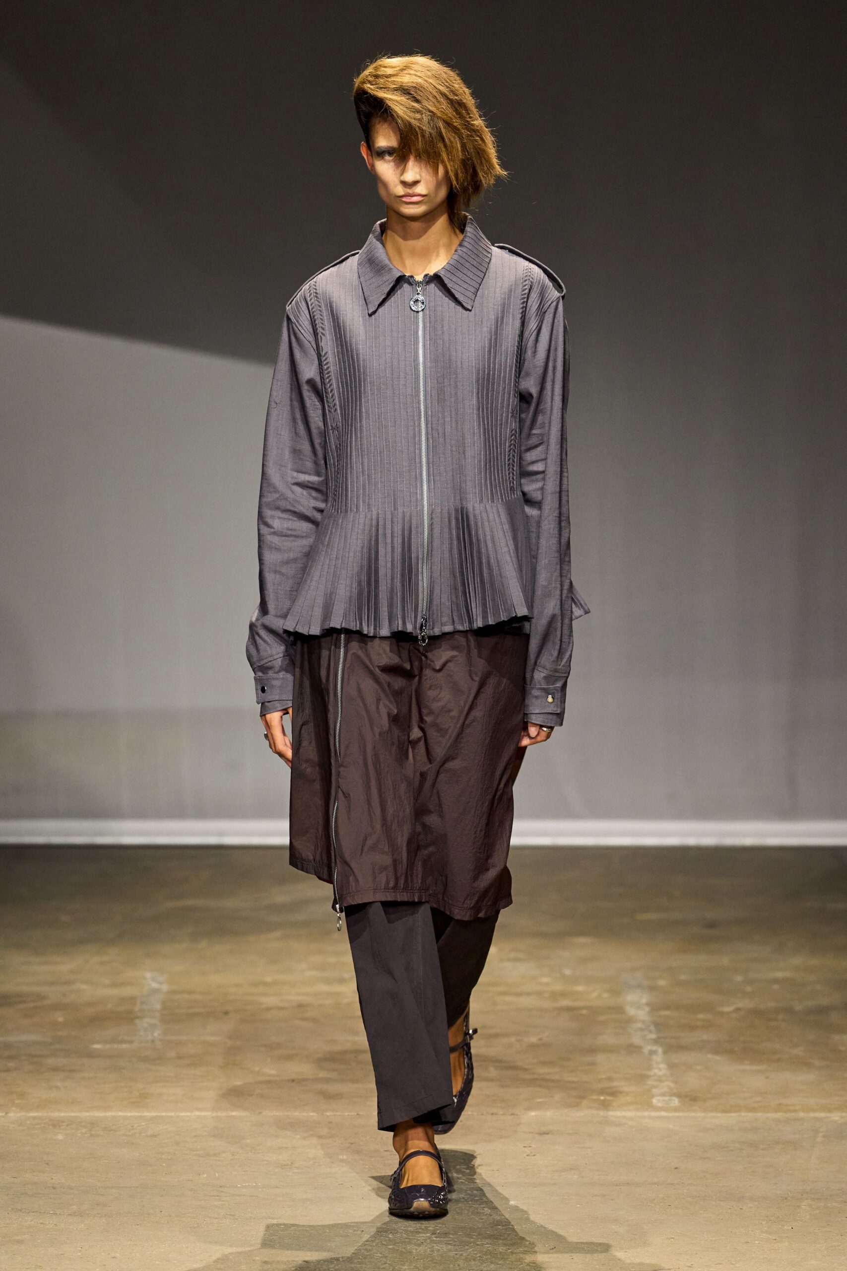 Slate zip-front jacket with knife pleats falling into a peplum hem. Deep brown crinkled skirt, off-center zip running nearly to the hem. Dark straight trousers peeking below the skirt. Mesh-toe Mary Jane flats with narrow straps. First train after rain, platform concrete still holding the damp.