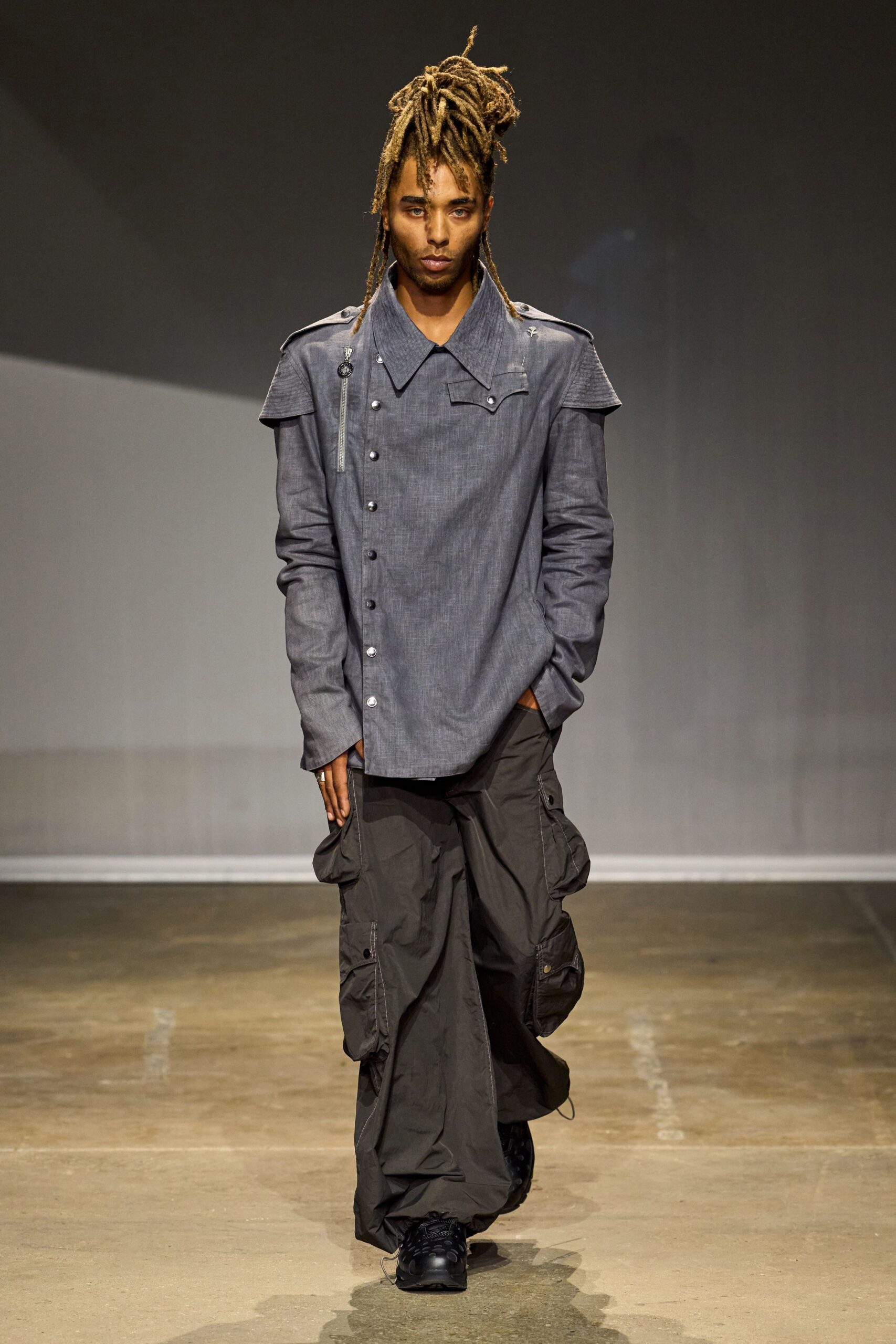 Steel chambray shirt with an off-center snap placket and zipped shoulder tab. Soot nylon cargos ballooning wide, flap pockets stacked down both legs. Black trail sneakers with thick laces. For crossing an empty terminal after the red-eye already landed.