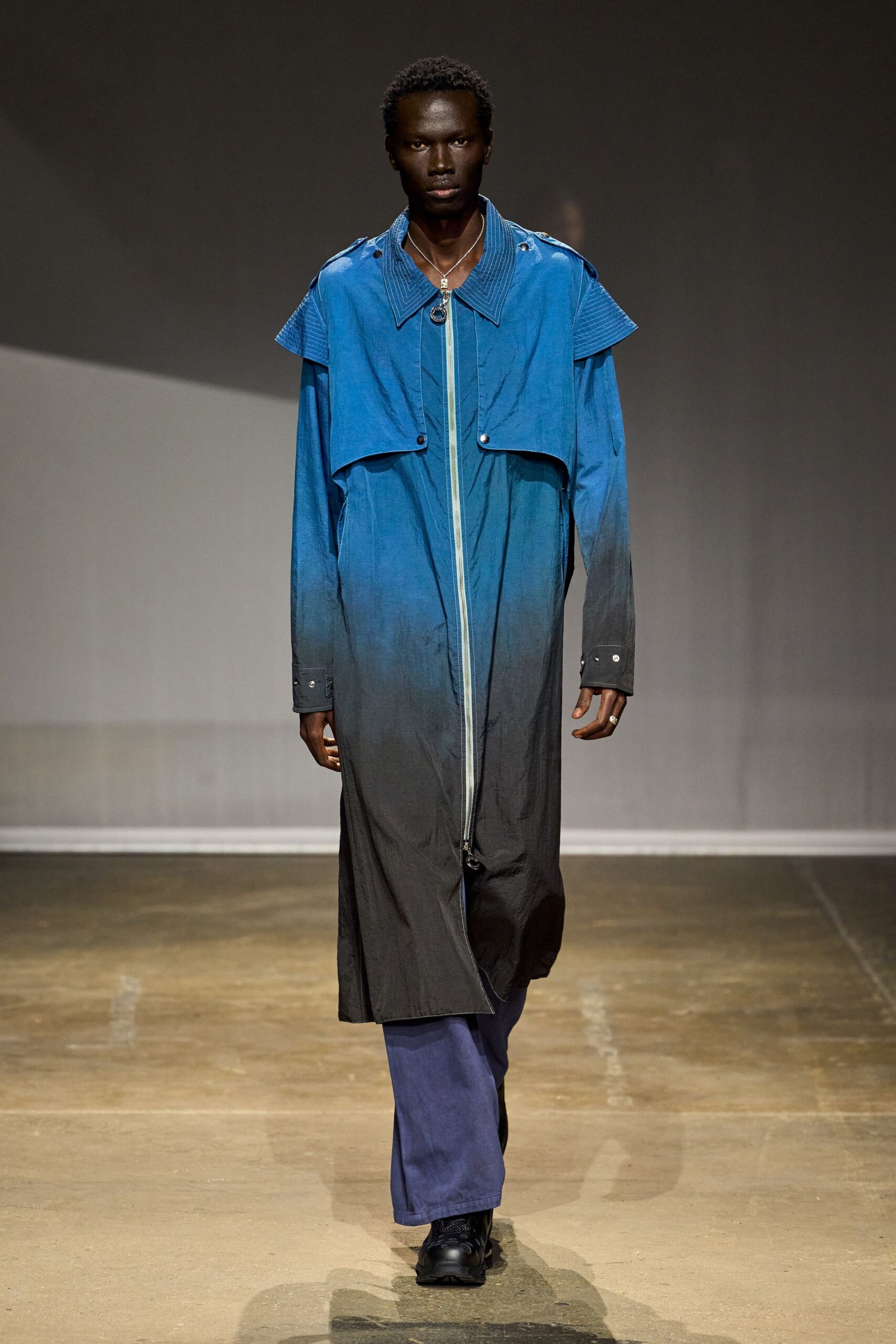Fine silver chain with a small square pendant. Cobalt to soot ombre coat in crinkled nylon, double cape yoke and long ring-pull zip. Indigo wide-leg trousers breaking over the shoe. Black trail shoes with thick rubber tread. Silver ring on one finger. Crossing an empty ferry terminal before the weather turns.