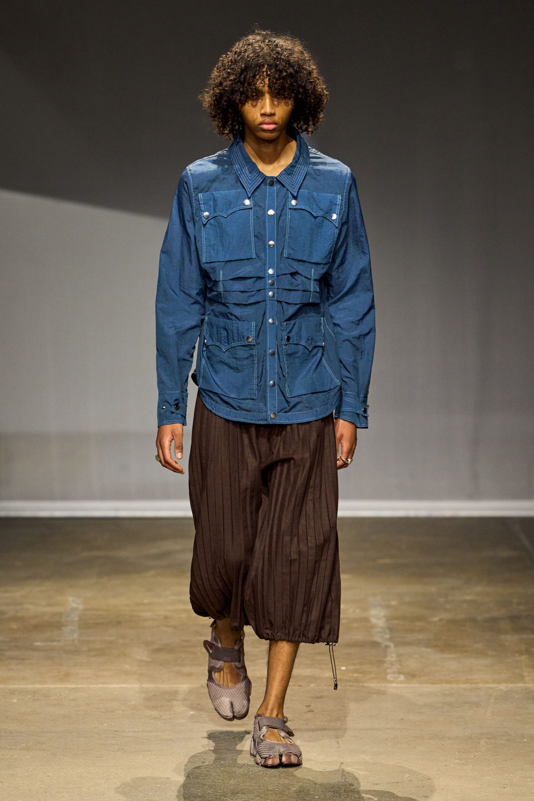 Indigo crinkled field jacket with white topstitching and silver snaps. Chocolate pinstriped harem trousers, gathered tight at the calf with a dangling cord end. Taupe mesh split-toe shoes strapped across the instep. Walking the long way home after rain, cuffs still damp from the curb.