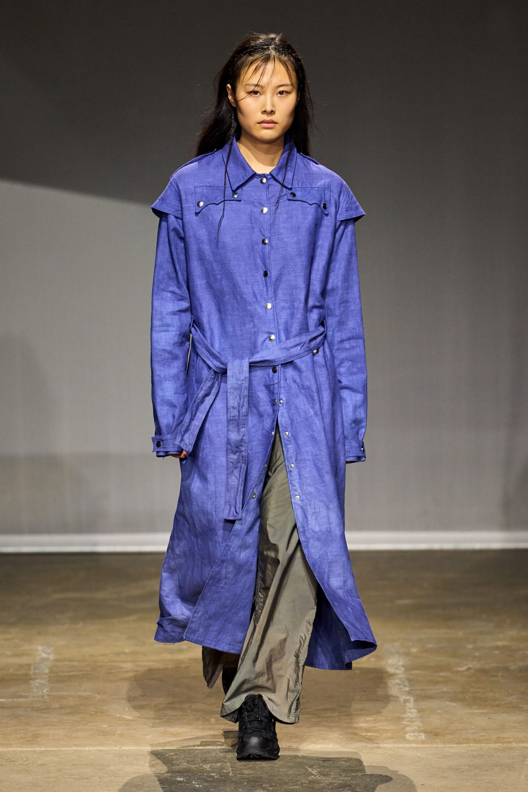 Cobalt linen coat with capelet shoulders and a self-tie hanging loose. Slate nylon wide-legs, crisp creases breaking over the shoe. Black trail sneakers with thick tread. Walking back from the ferry in a wind that keeps changing sides.