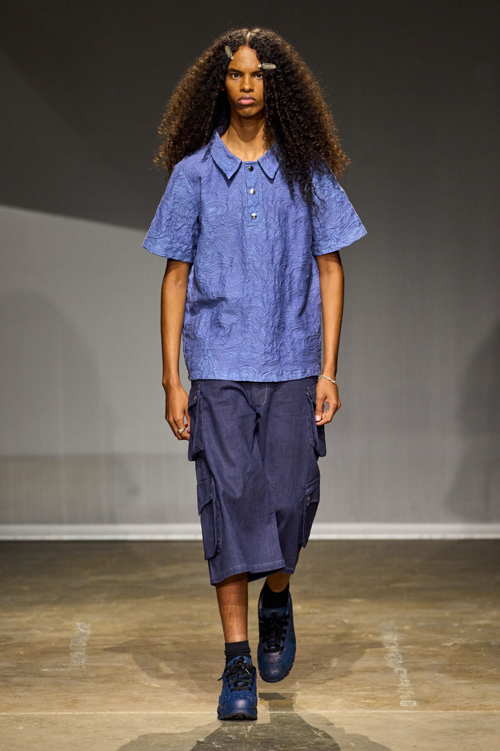 Periwinkle jacquard polo shirt traced with woodgrain swirls and a three-snap placket. Ink blue knee-length cargo shorts in slubby twill, flap pockets stacked at the thigh. Black ankle socks. Cobalt lace-up clogs punched with round ventilation holes. Walking across a hot concrete quad before the summer workshop starts.