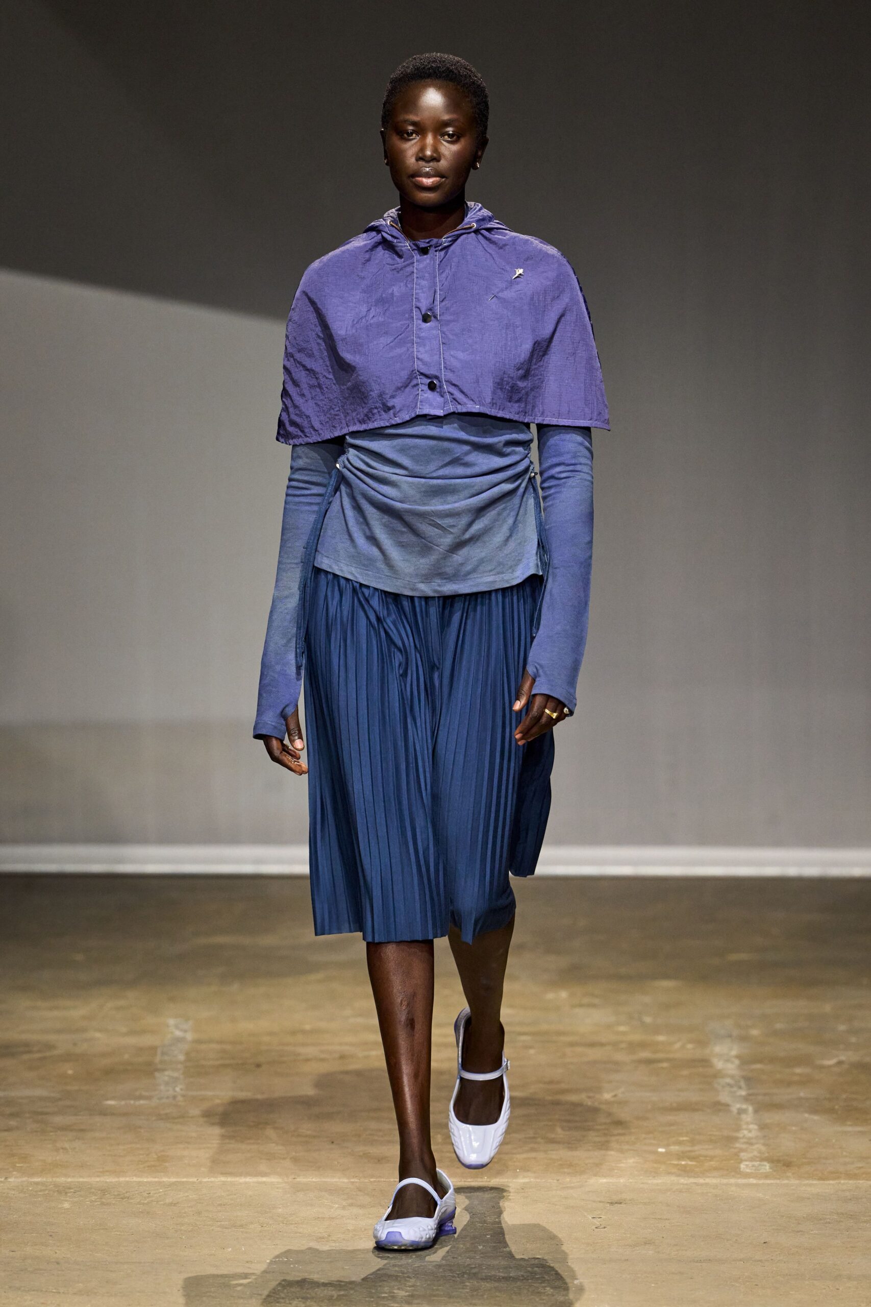 Crinkled violet capelet with black buttons and a tiny chest pin. Washed blue jersey wrapped close, drawstrings hanging loose at both hips. Ink-blue pleated midi, the front split opening with each step. Pale lavender Mary Jane flats with translucent purple soles. Walking the aquarium tunnel after school lets out.