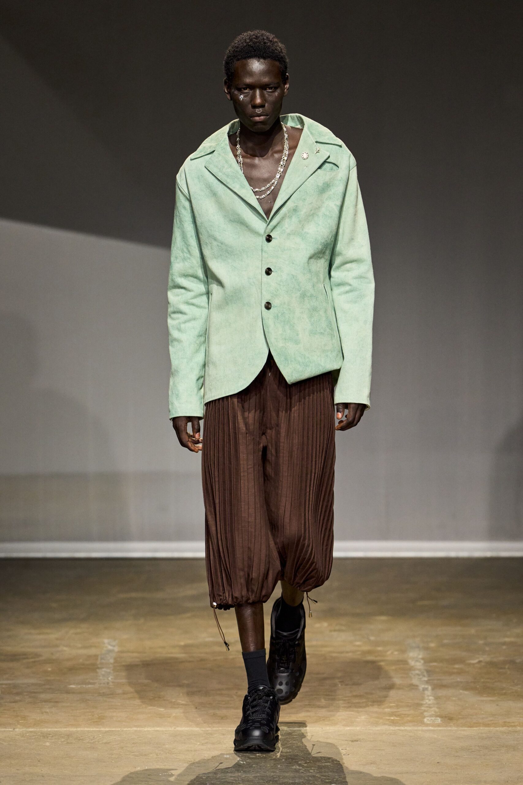 Layered silver chains dropping into the open chest. Mint suede jacket, clouded finish and off-center collar pin. Chocolate pleated bloomers cinched at the calf with dangling cords. Black trainers with perforated toes and thick rope laces. Black ankle socks. Crossing the conservatory after watering, concrete still damp underfoot.