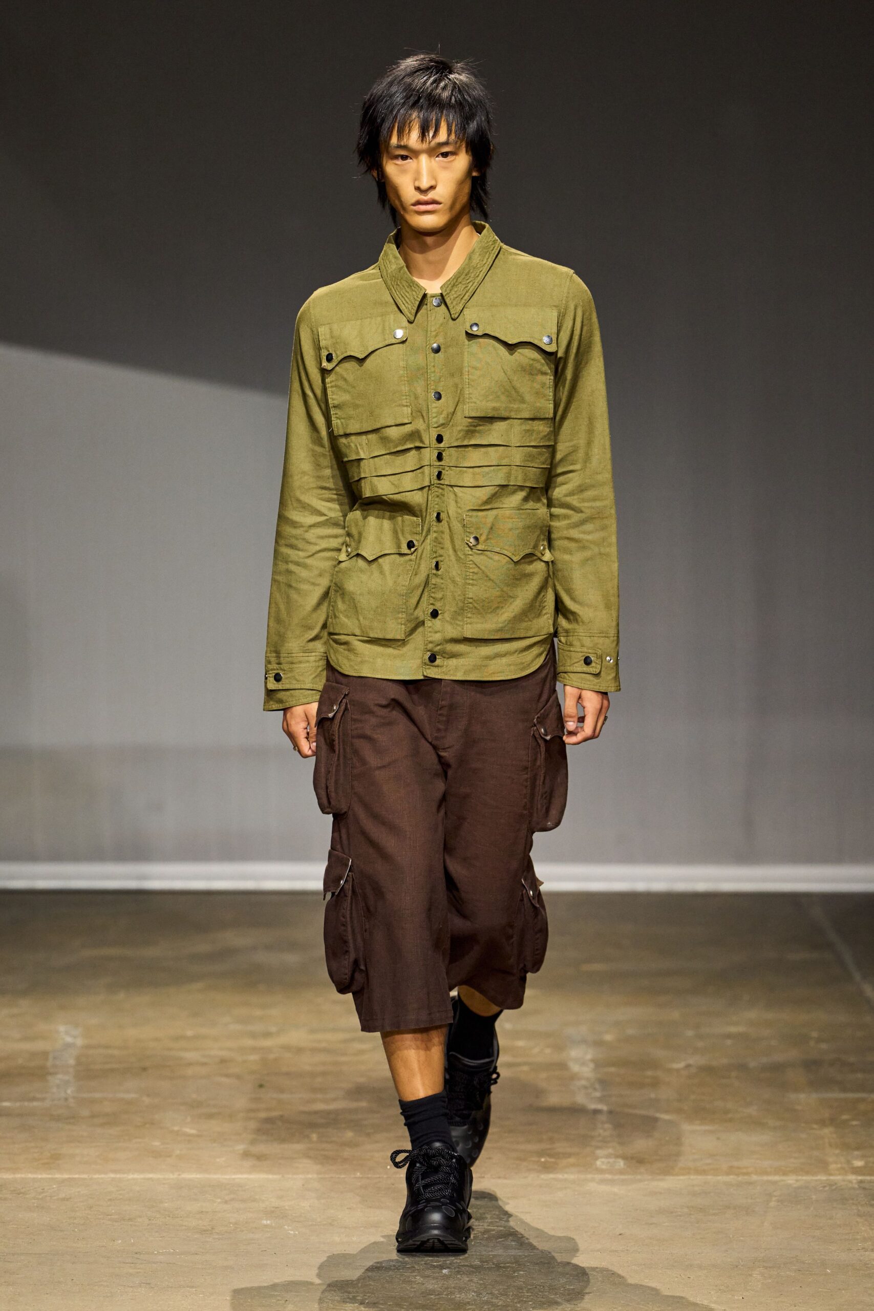 Olive cotton field jacket, pin-tucked through the ribs with black snaps. Chocolate cargo shorts cut long, bellows pockets swinging at both thighs. Black crew socks scrunched at the ankle. Black lace-up shoes with padded collars. For checking the irrigation lines before the heat starts up.
