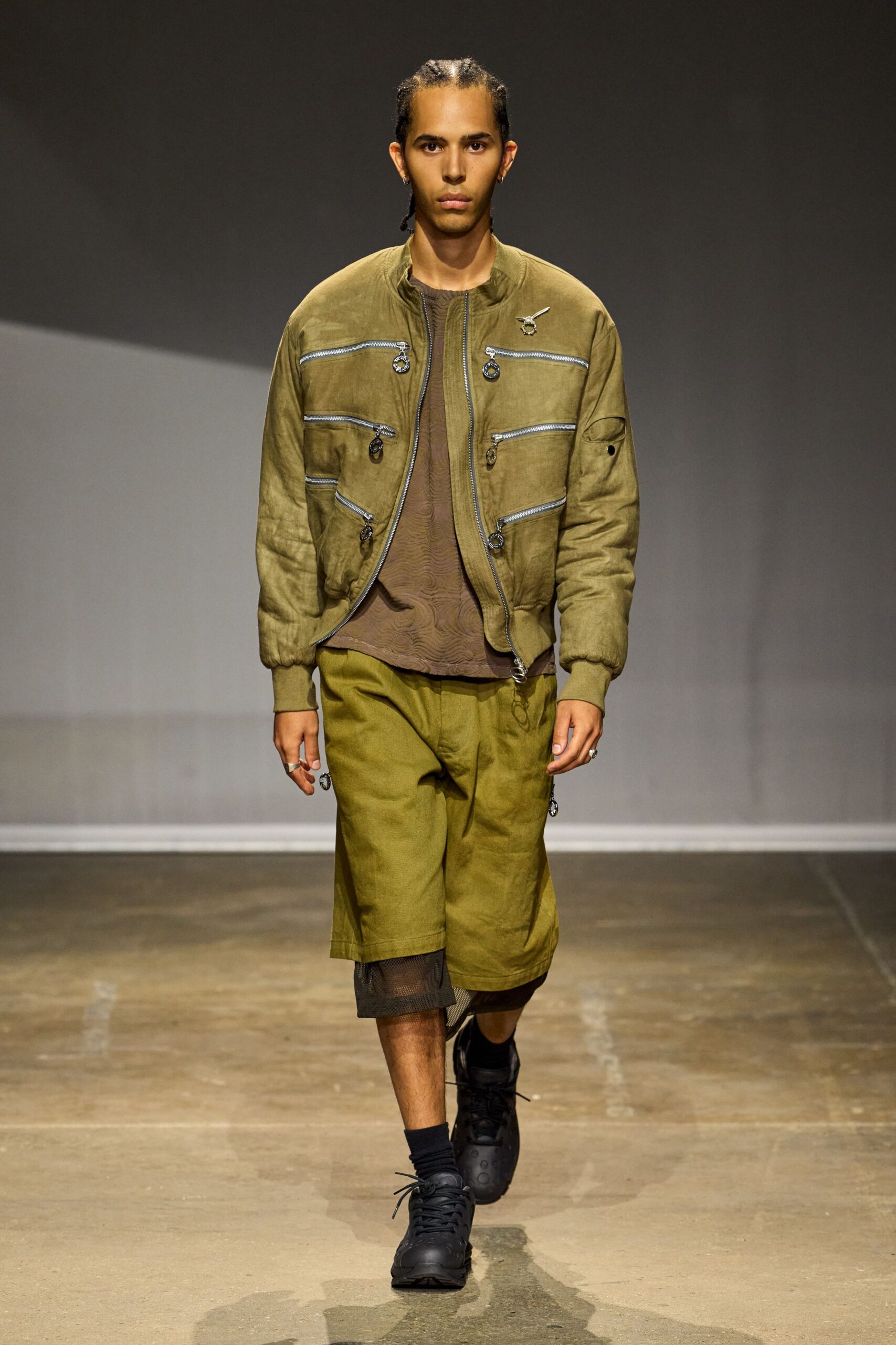 Olive bomber crowded with horizontal zips and ring pulls. Sheer brown mesh top with moire ripples across the chest. Moss knee-length shorts in washed twill. Black mesh panel hanging below one hem. Bulky black sneakers with punched rubber toe caps. For crossing an airfield runway in heat that warps the distance.