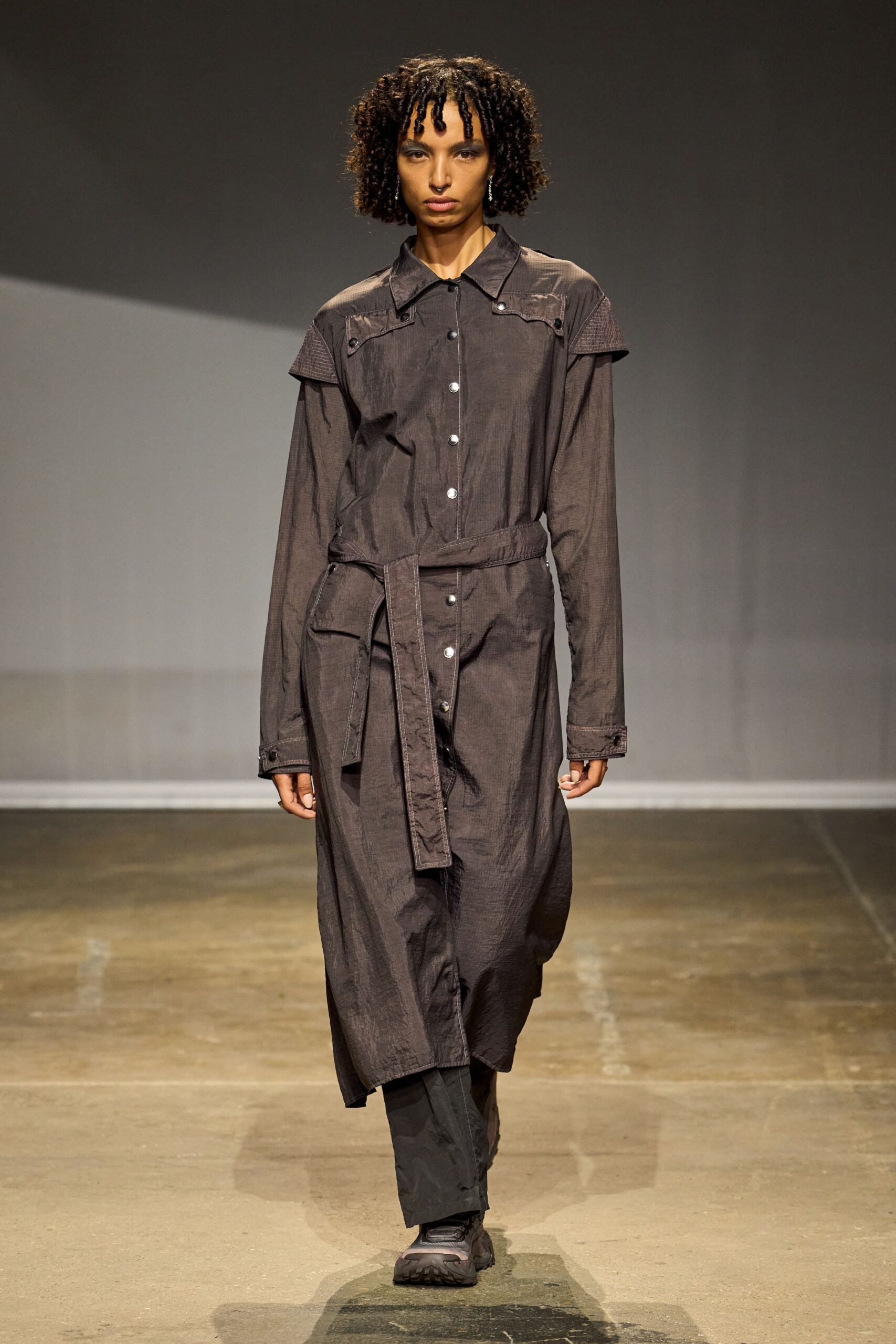 Charcoal crinkled shirt dress with snap placket, epaulets, and a long self belt. Black straight trousers peeking below the uneven hem. Black trail shoes with ridged rubber soles. For crossing an empty transit terminal before the first train starts.
