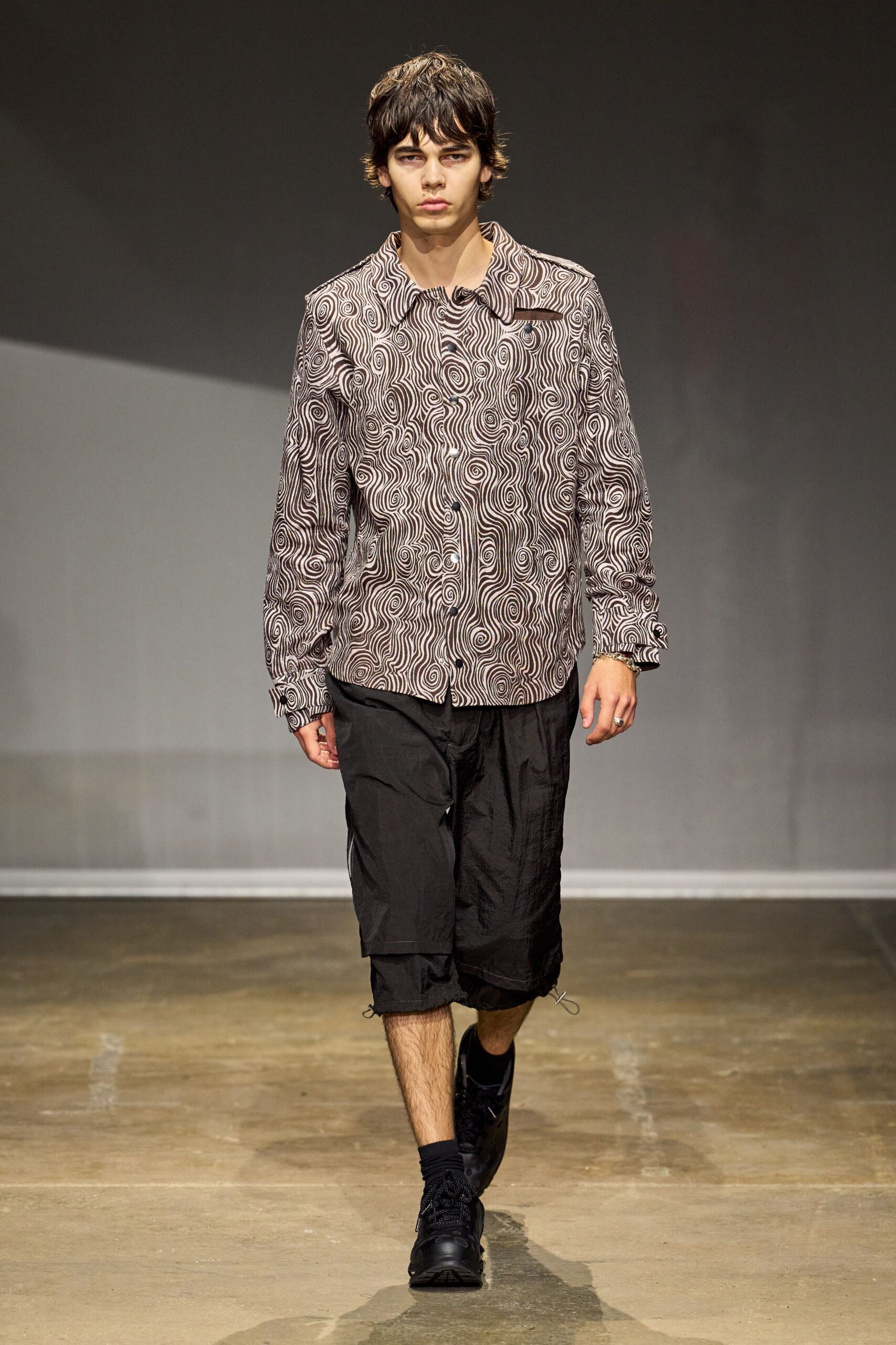 Brown-and-cream swirl print shirt, oversized collar and a slit chest welt. Black crinkled nylon long shorts, raw hem grazing the shin. Black low sneakers with thick rope laces. Black ankle socks scrunched soft at the cuff. Walking out of a print studio with fresh ink still under one nail.